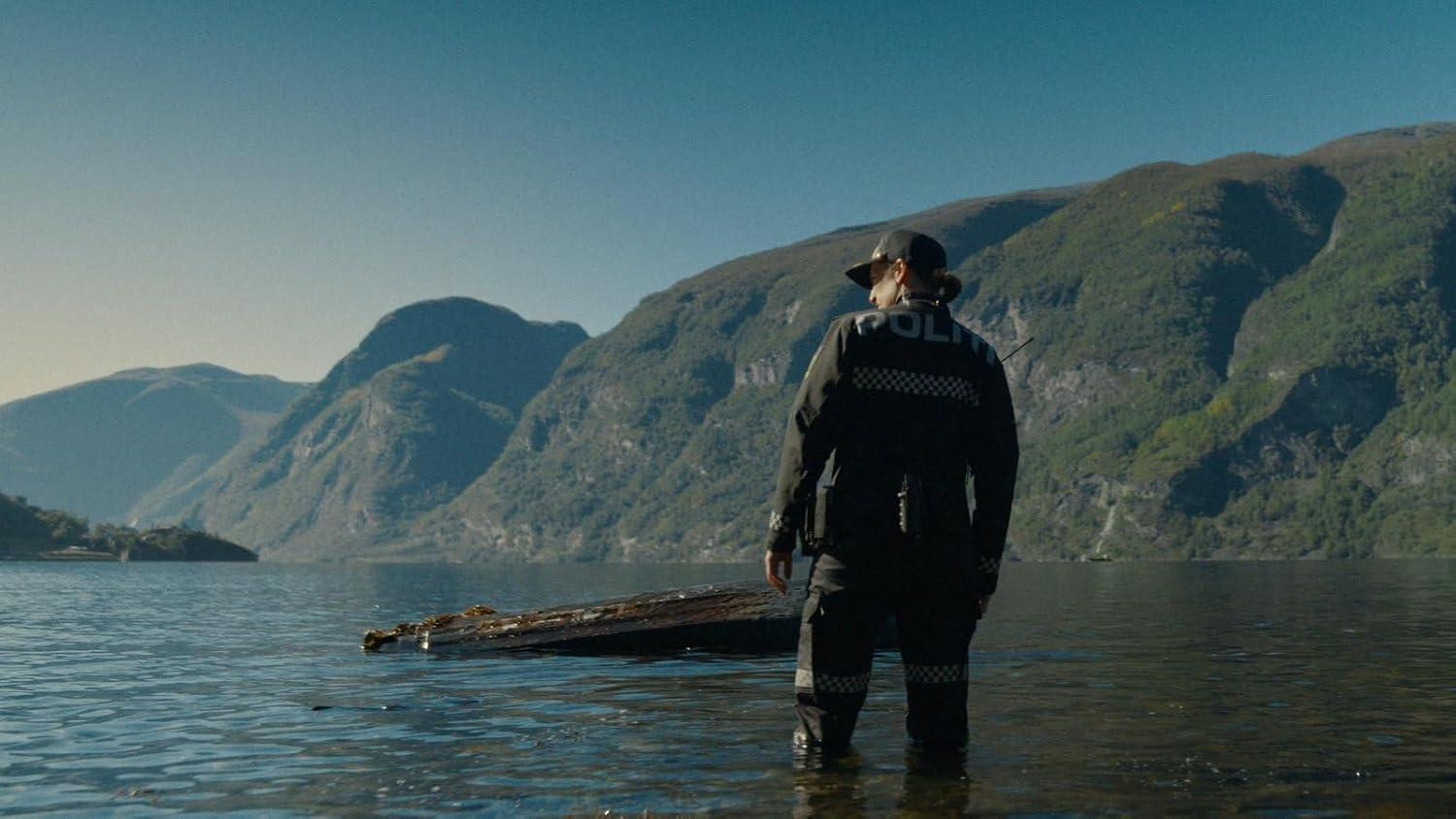 Ein Polizeibeamter in Uniform steht im flachen Wasser eines Fjords vor einer bergigen Kulisse.