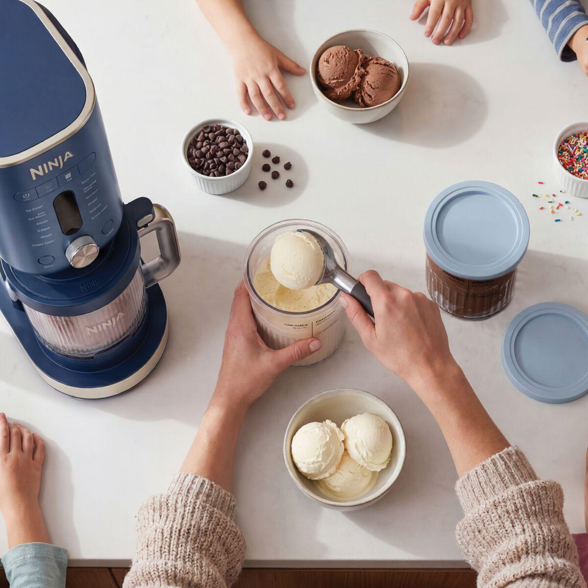 Person schöpft Vanilleeis an einer Arbeitsplatte mit Eismaschine, Toppings und Schüsseln.