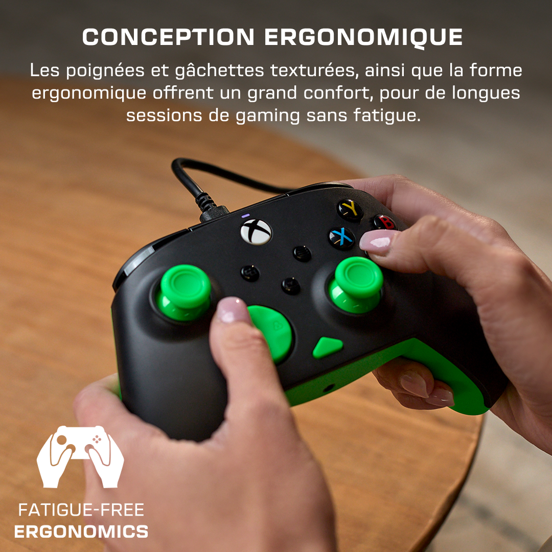 Une personne tient une manette Xbox noire avec des joysticks et des accents vert vif sur une table en bois.