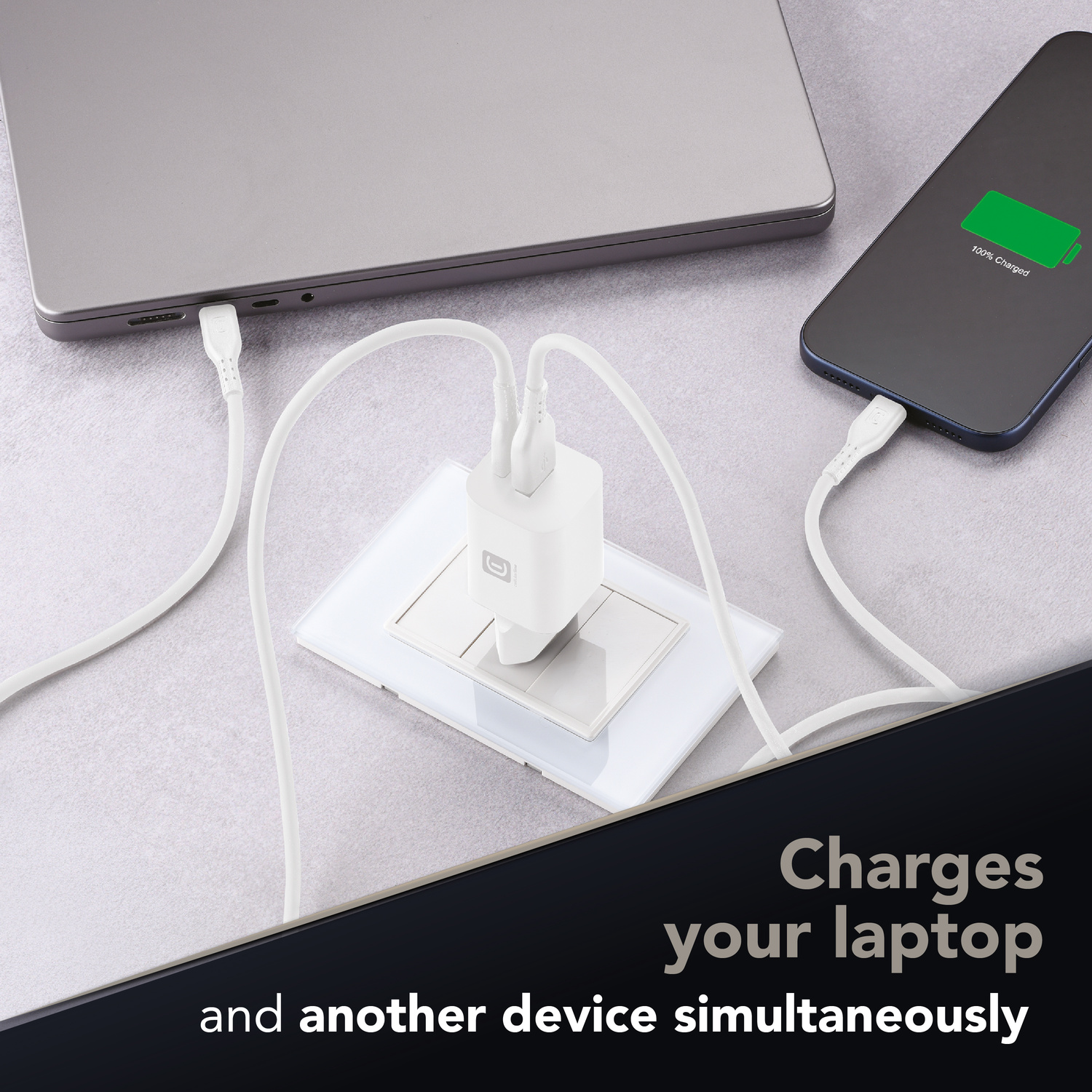 Un caricatore da muro USB bianco carica contemporaneamente un computer portatile e uno smartphone su una superficie grigia.