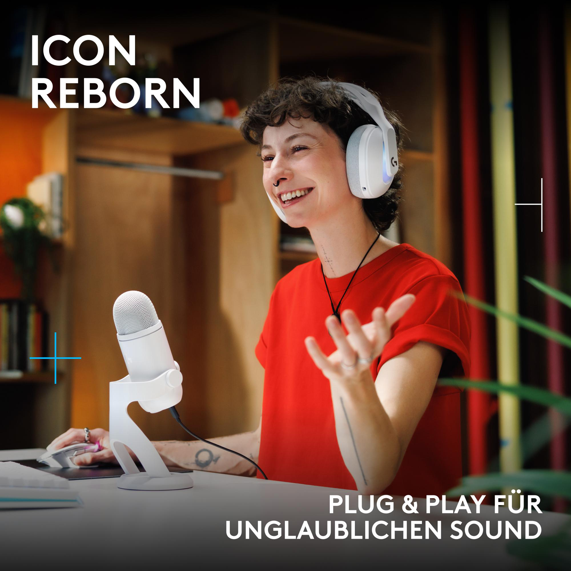 Eine lächelnde Person mit rotem Shirt und weißem Headset spricht in ein weißes Mikrofon vor einem Schreibtisch.
