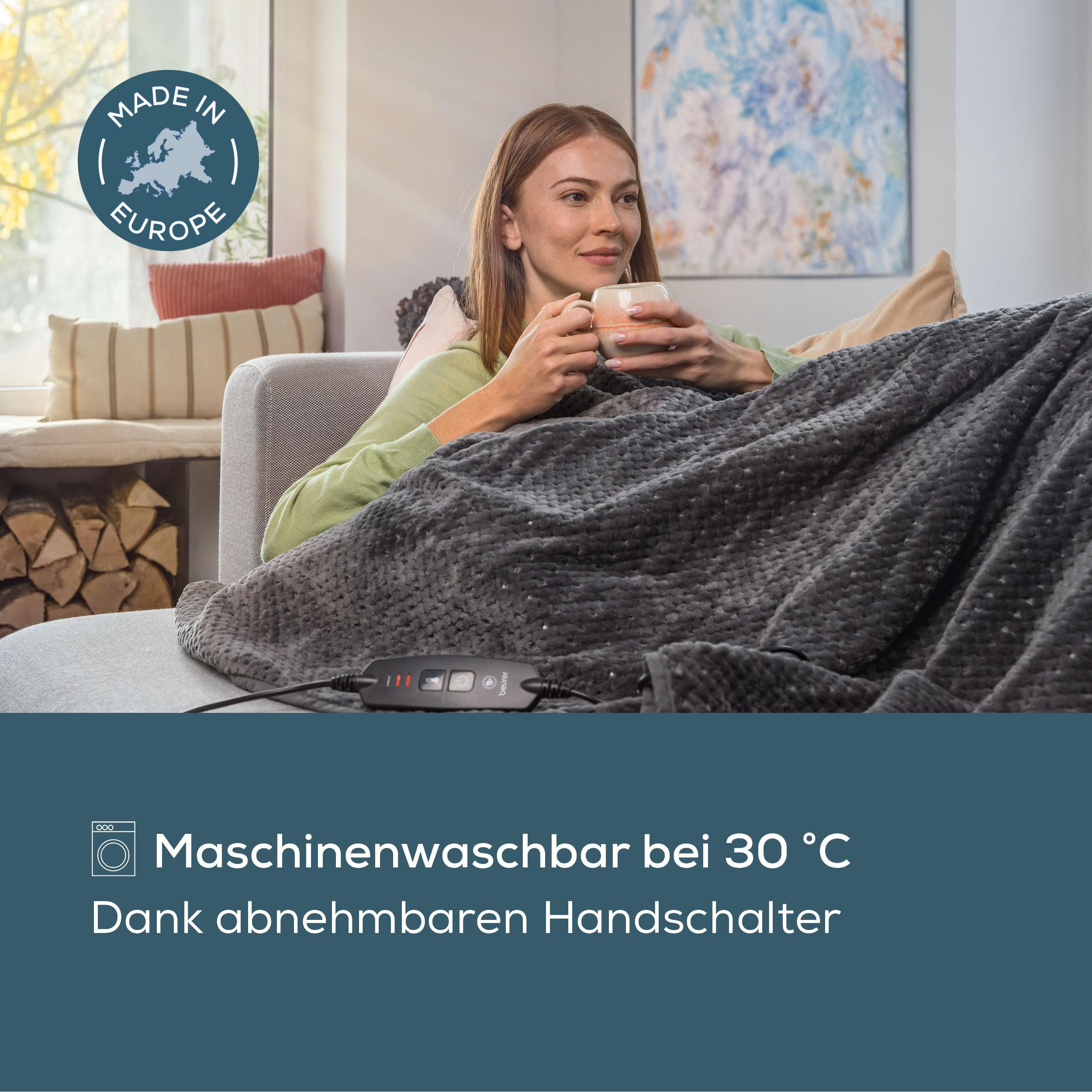 Frau sitzt gemütlich unter einer grauen Heizdecke auf dem Sofa, hält eine Tasse und hält den Handschalter.
