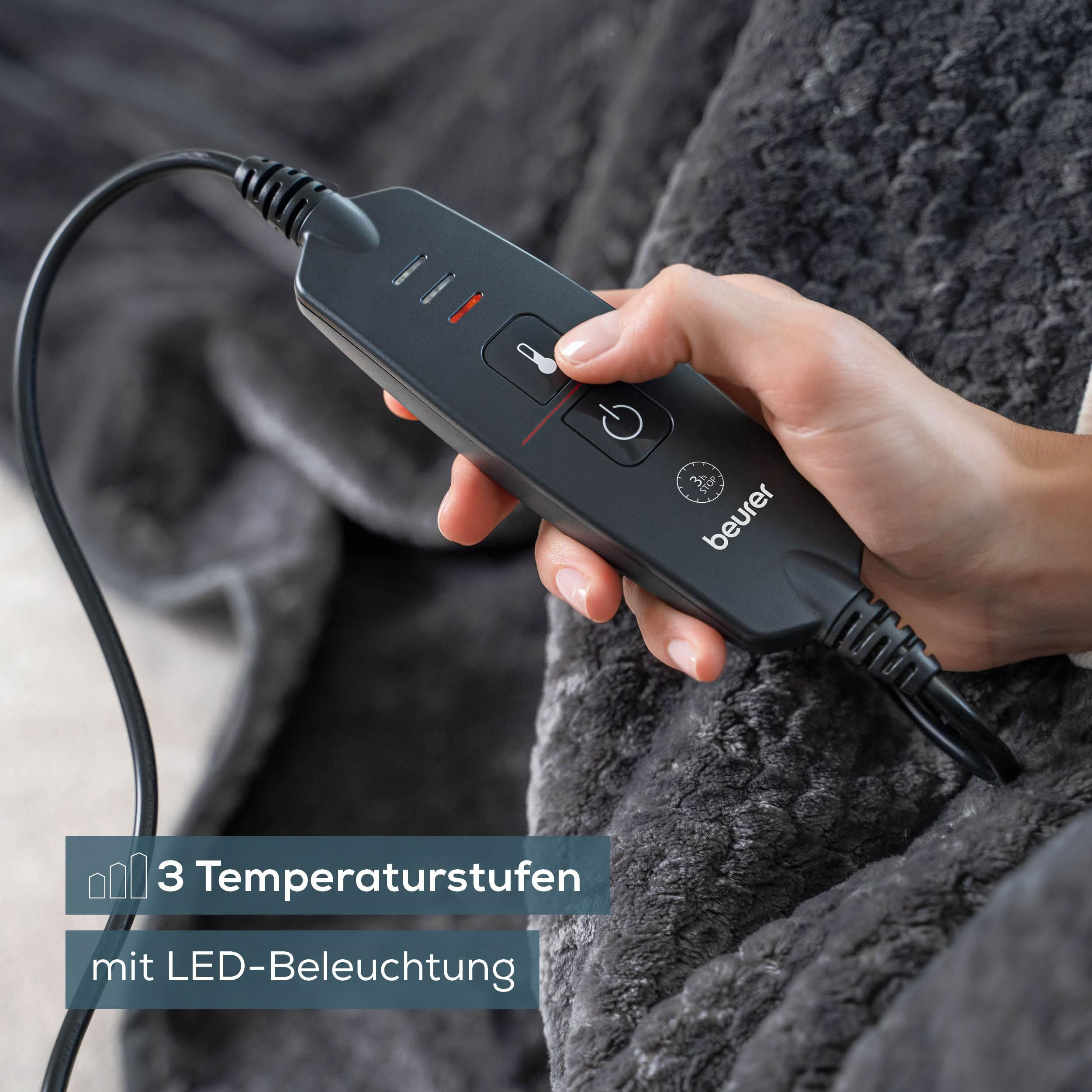 Hand hält eine schwarze Beurer-Fernbedienung mit drei Temperaturstufen und LED-Beleuchtung vor einer grauen Decke.