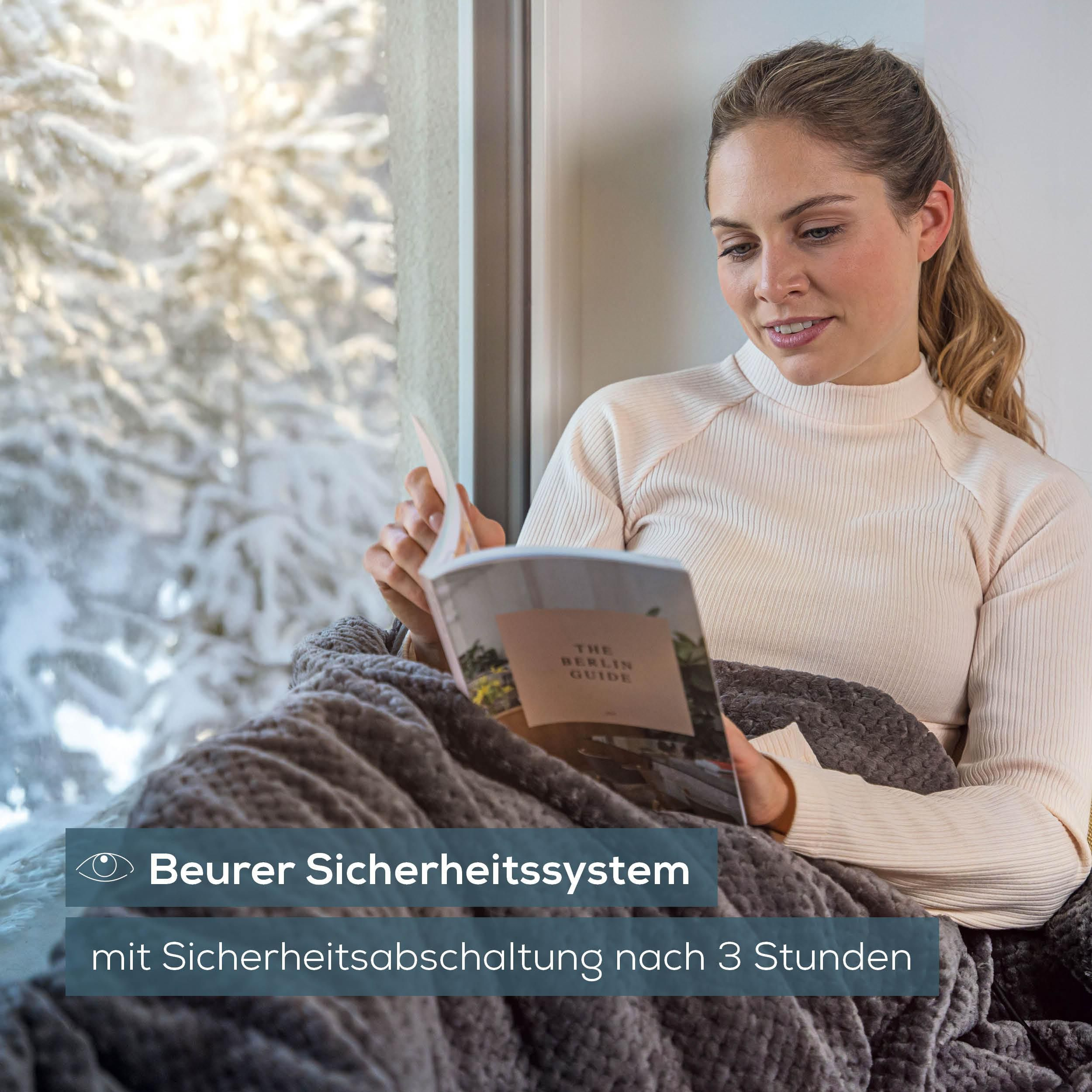 Eine blonde Frau sitzt eingekuschelt in eine graue Decke am Fenster und liest ein Berlin-Reiseheft.