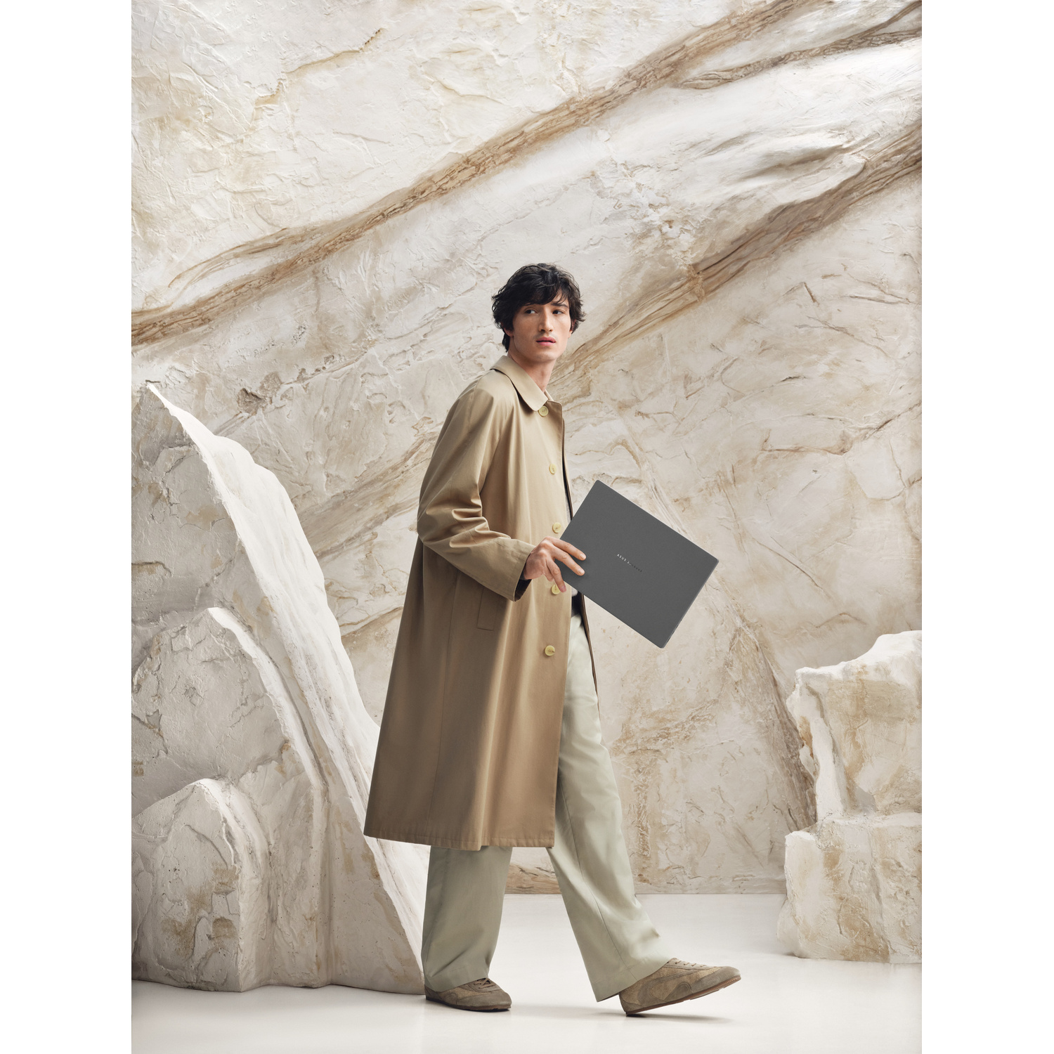 Un uomo con trench beige cammina davanti a una parete rocciosa testurizzata tenendo un laptop.