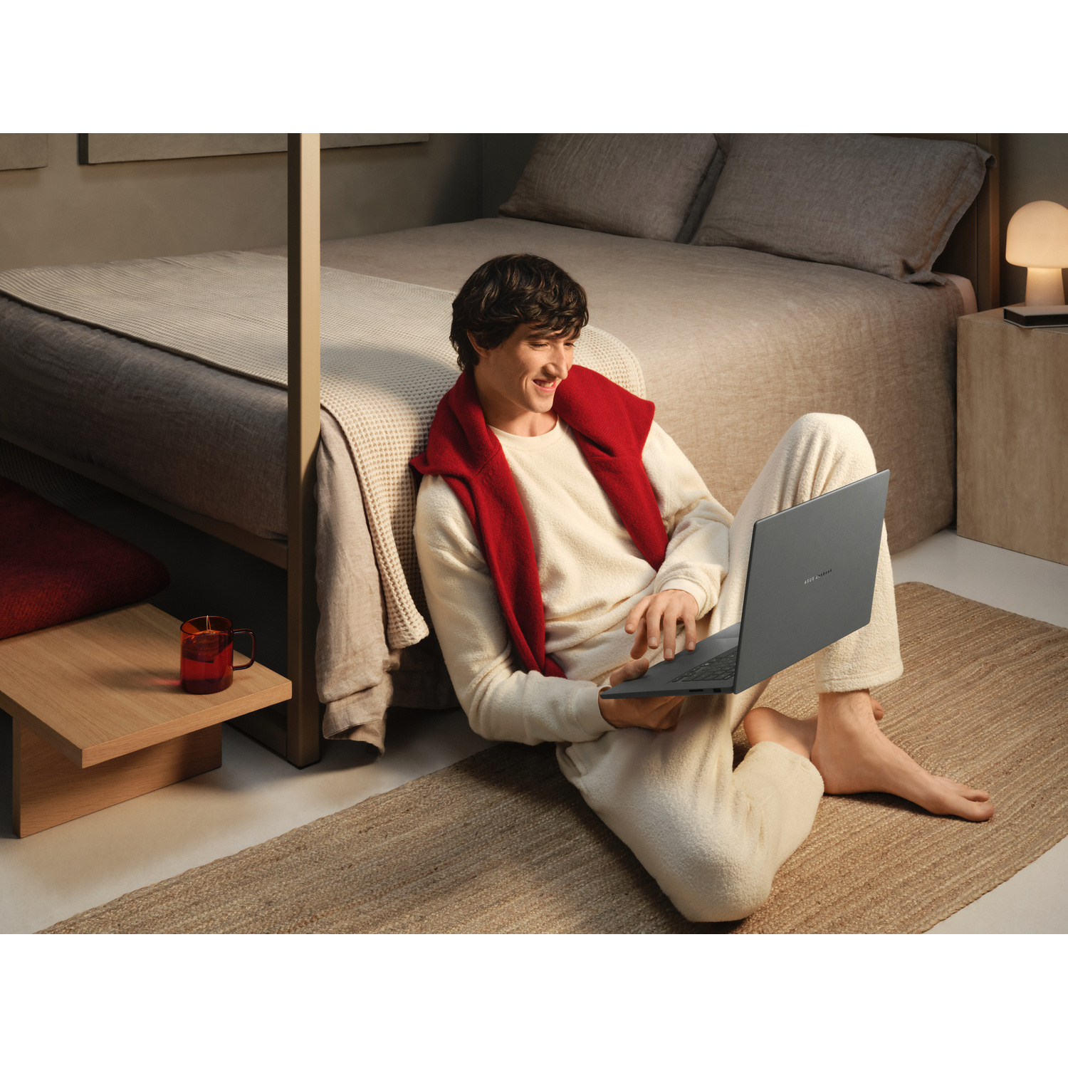 Un uomo in pigiama beige e maglione rosso siede sul tappeto della camera da letto usando un laptop.