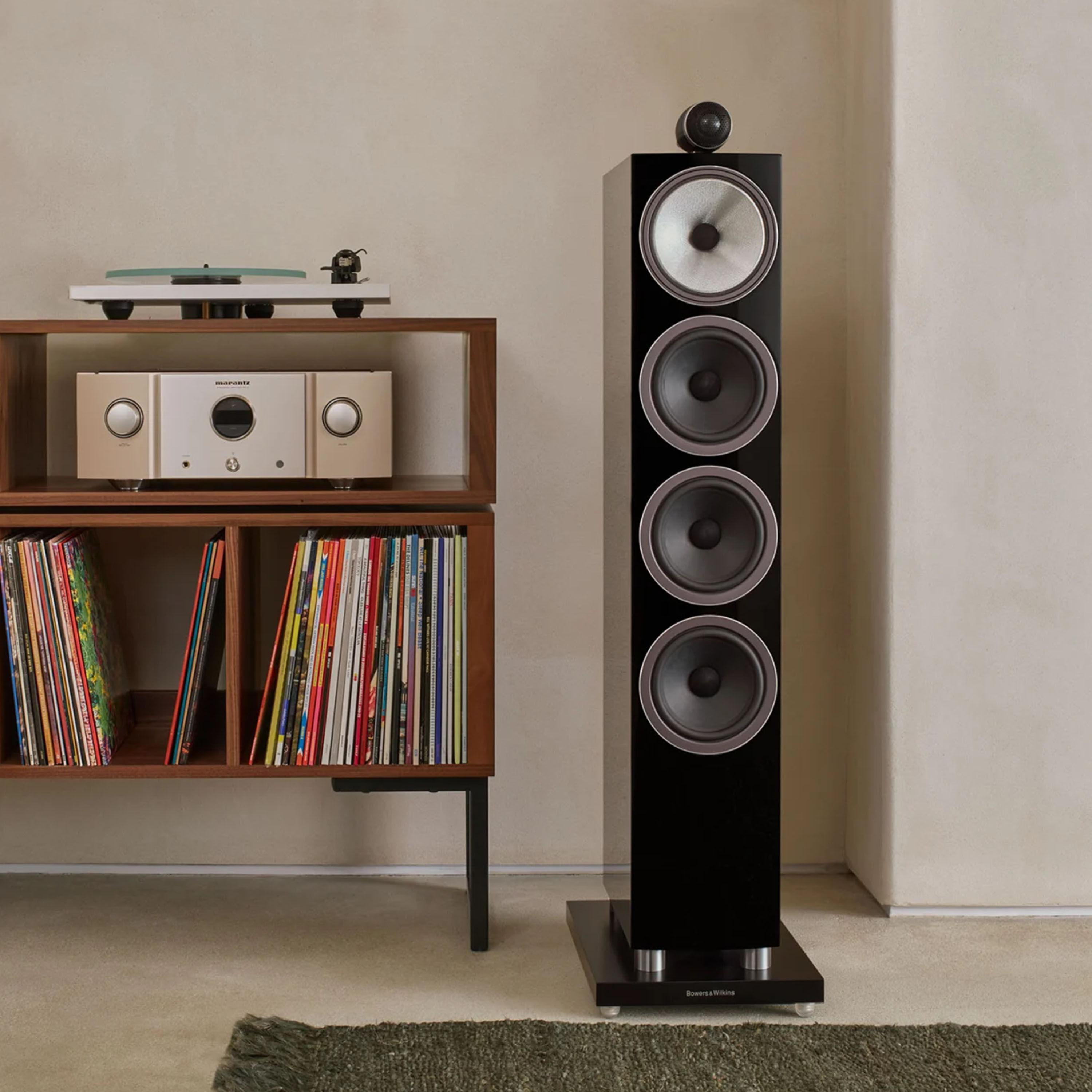 Ein hoher schwarzer Standlautsprecher steht neben einem Holzregal mit Receiver und Schallplatten.