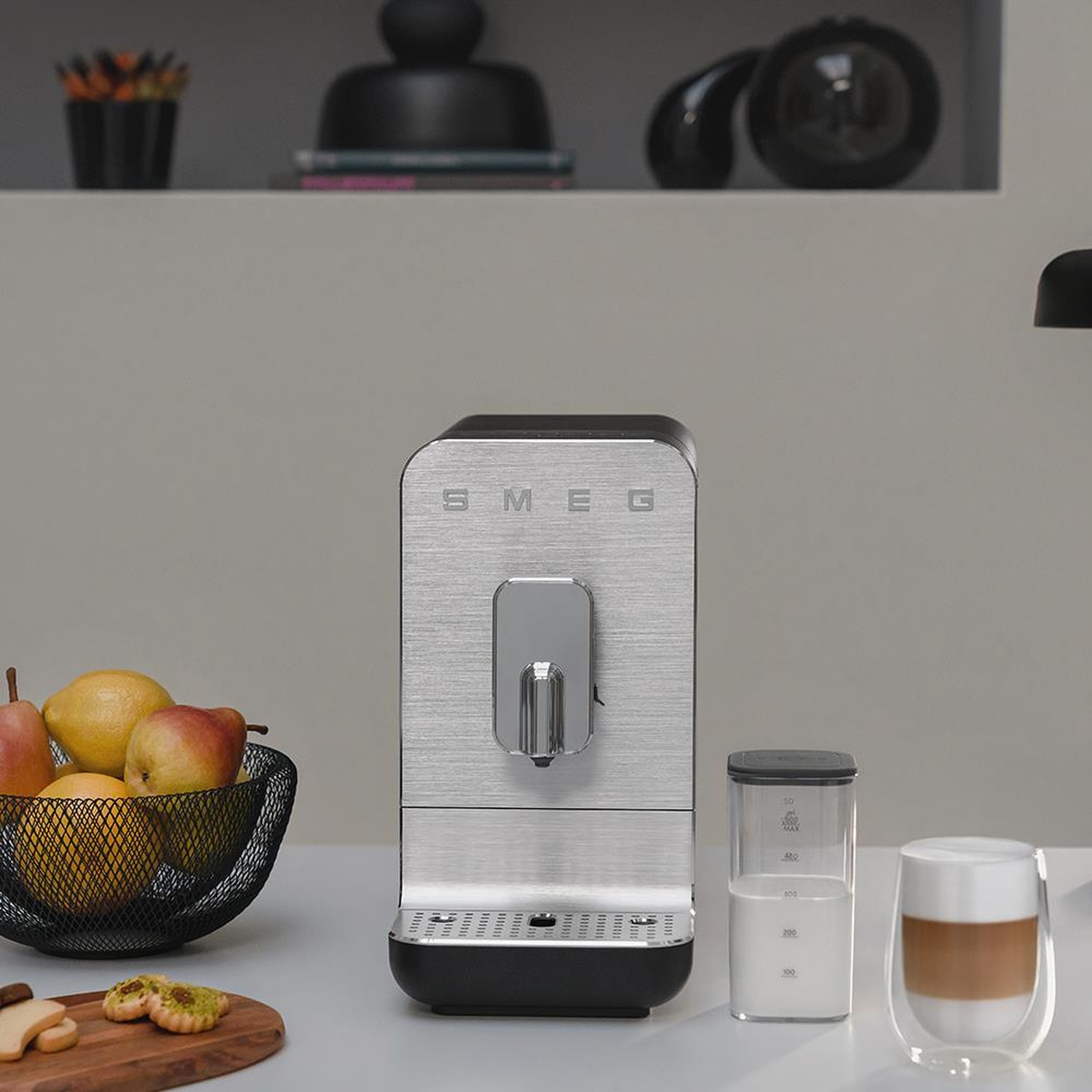 Una macchina da caffè Smeg argentata si trova su un bancone con frutta, latte e un cappuccino.