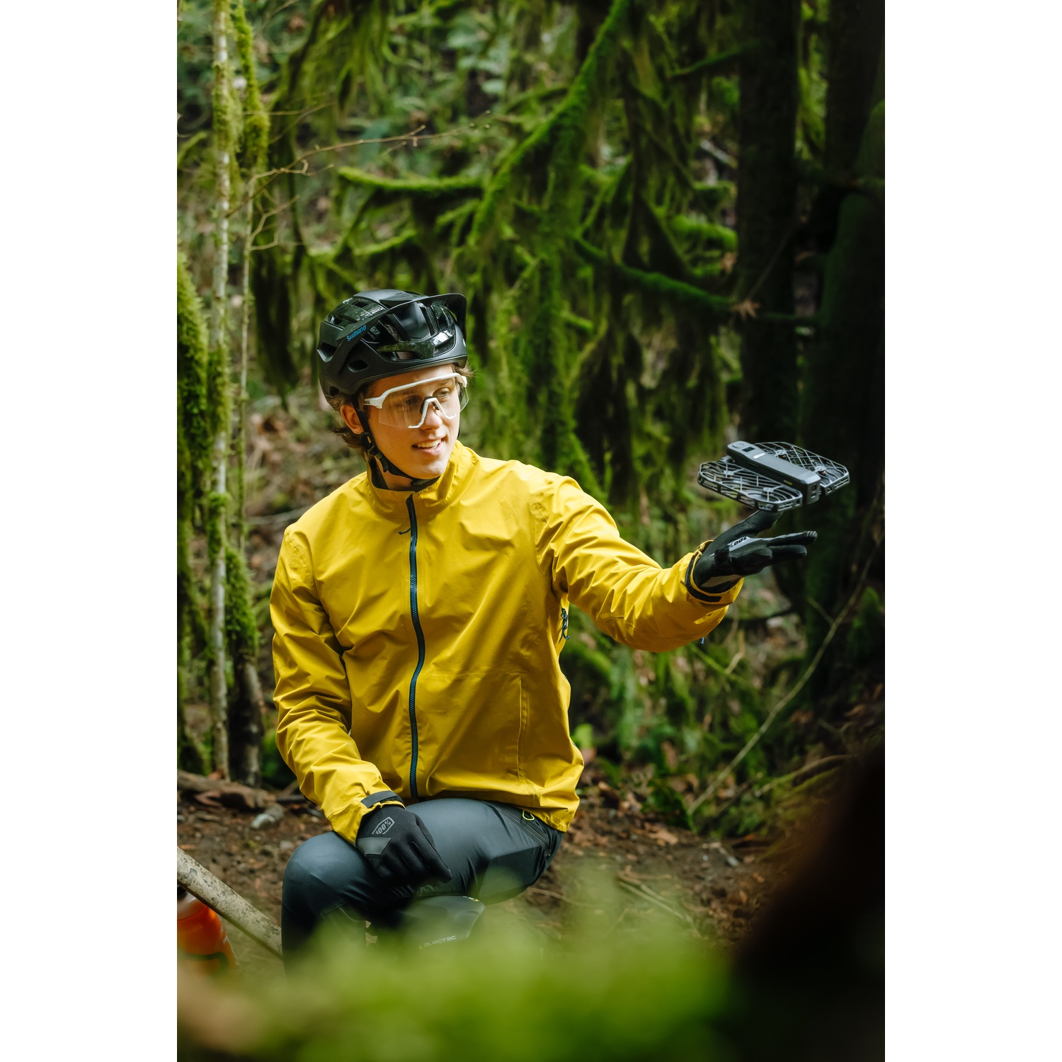 Un ciclista con giacca gialla e casco lancia un piccolo drone dalla mano in un bosco muschioso.
