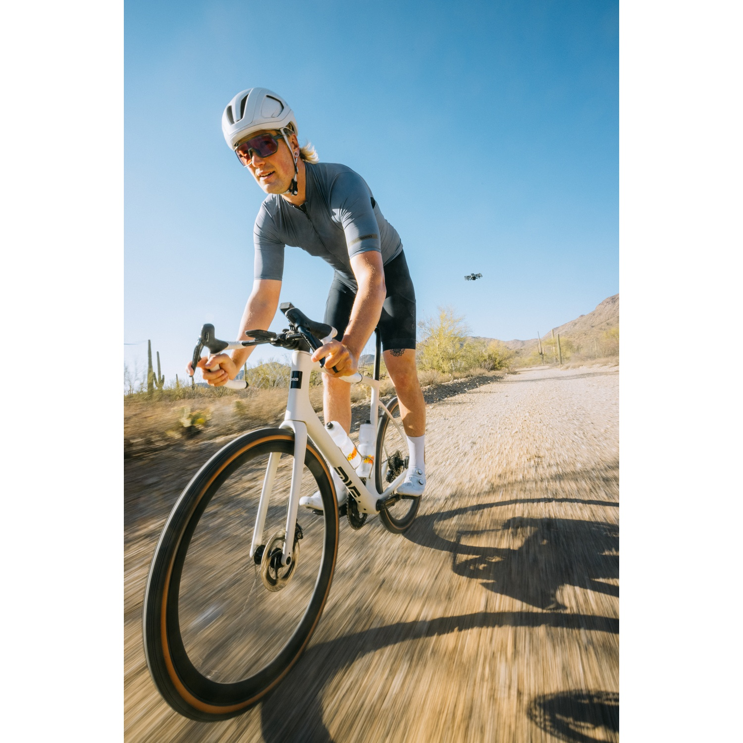 Un ciclista con maglia grigia e casco bianco pedala su una strada sterrata soleggiata nel deserto.