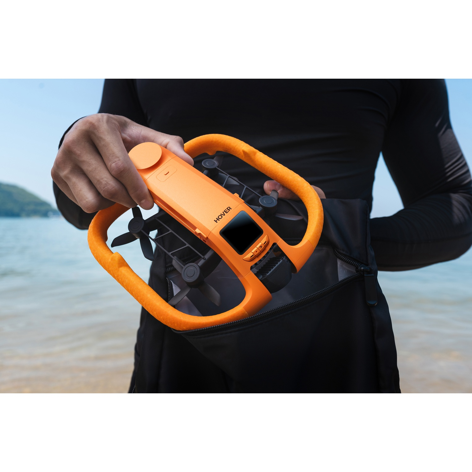 Una persona estrae un drone arancione da uno zaino nero con un lago sullo sfondo.