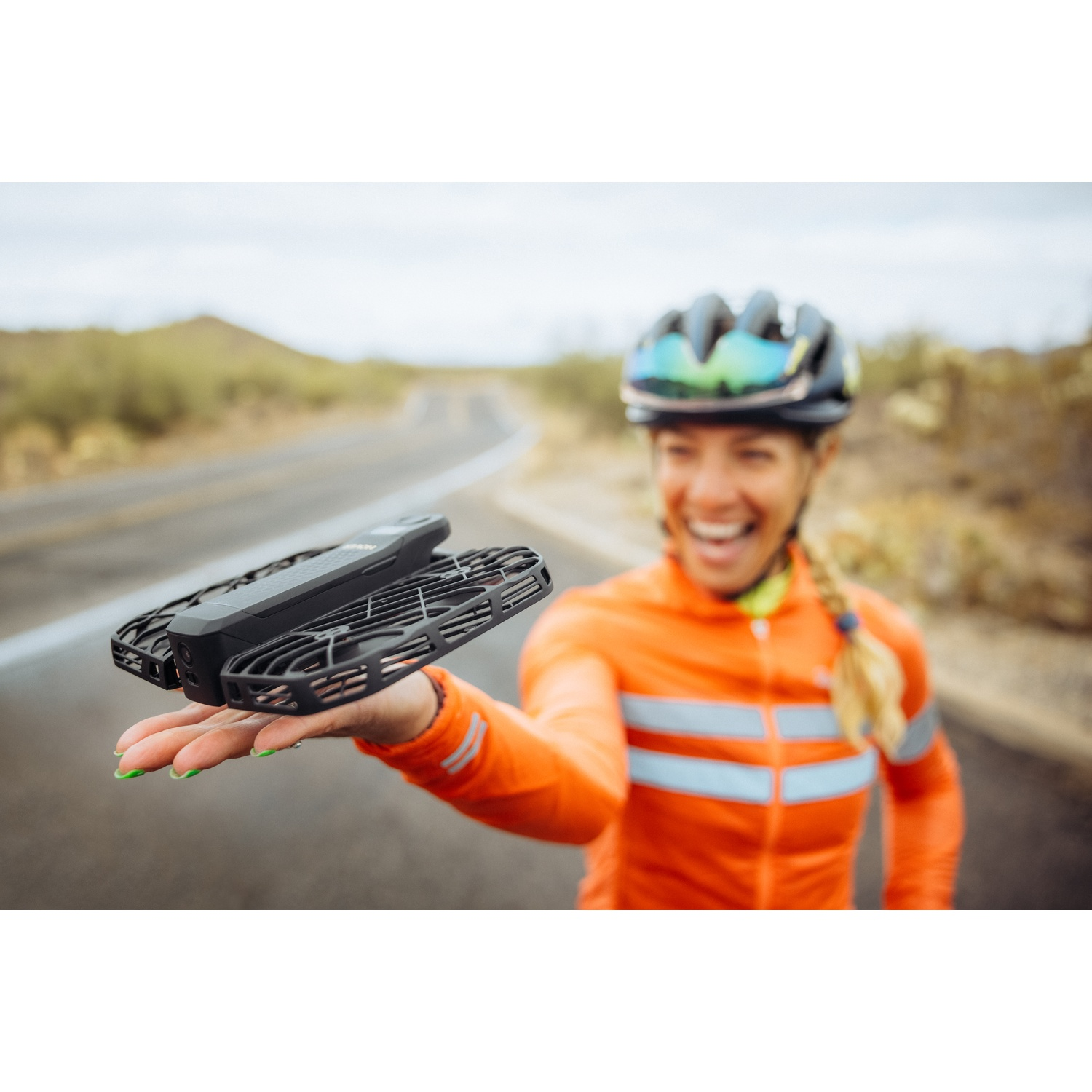 Una ciclista sorridente in giacca arancione tiene un drone nero in mano su una strada.