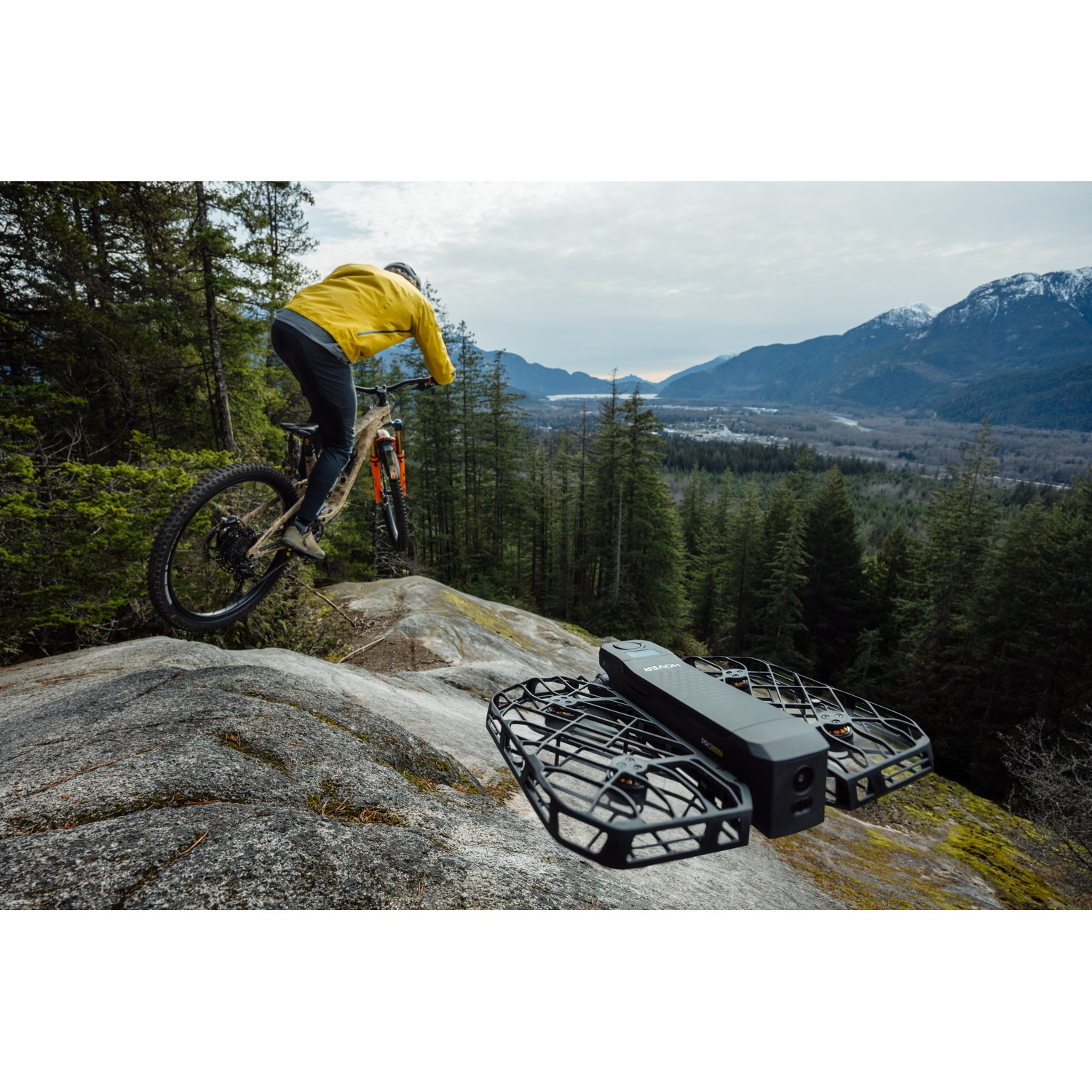 Un drone vola su una cresta rocciosa mentre un mountain biker passa nello sfondo con montagne.