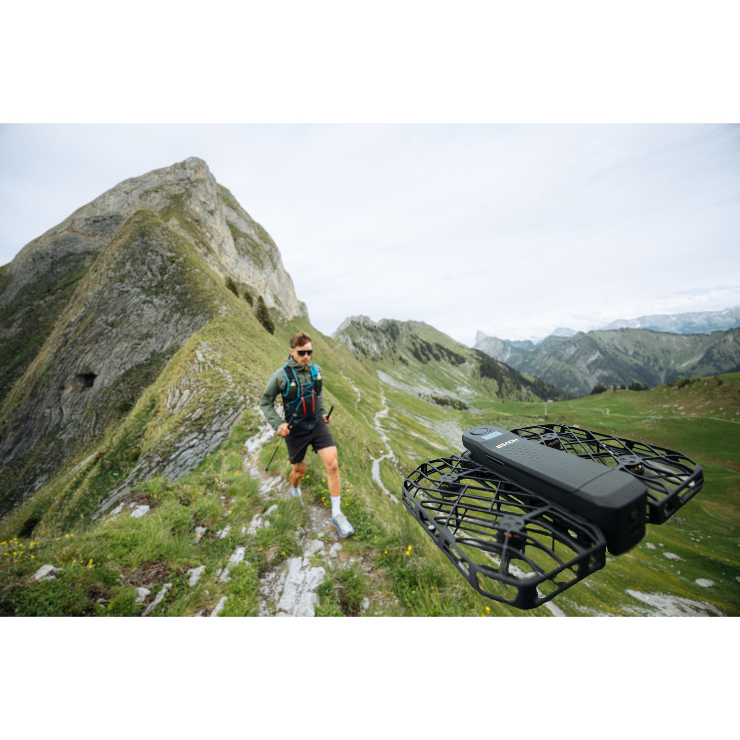 Un drone nero vola in aria mentre un escursionista cammina lungo un sentiero di montagna sullo sfondo.