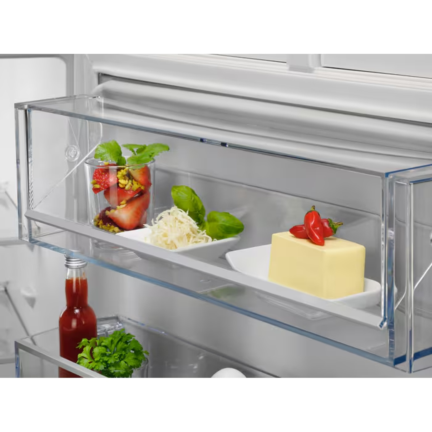 Open refrigerator door bin holding a glass of fruit, grated cheese with basil, and a block of butter with small peppers.