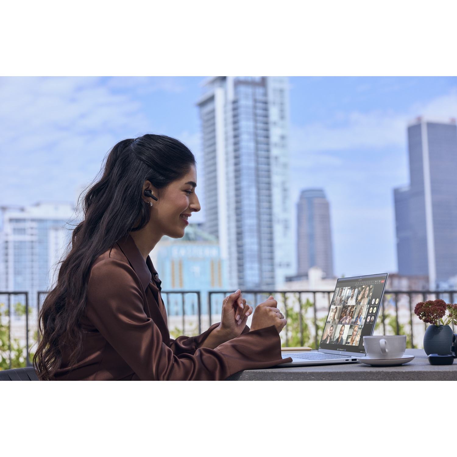 Een vrouw in een bruine blouse zit op een balkon en voert een videogesprek op haar laptop met skyline.