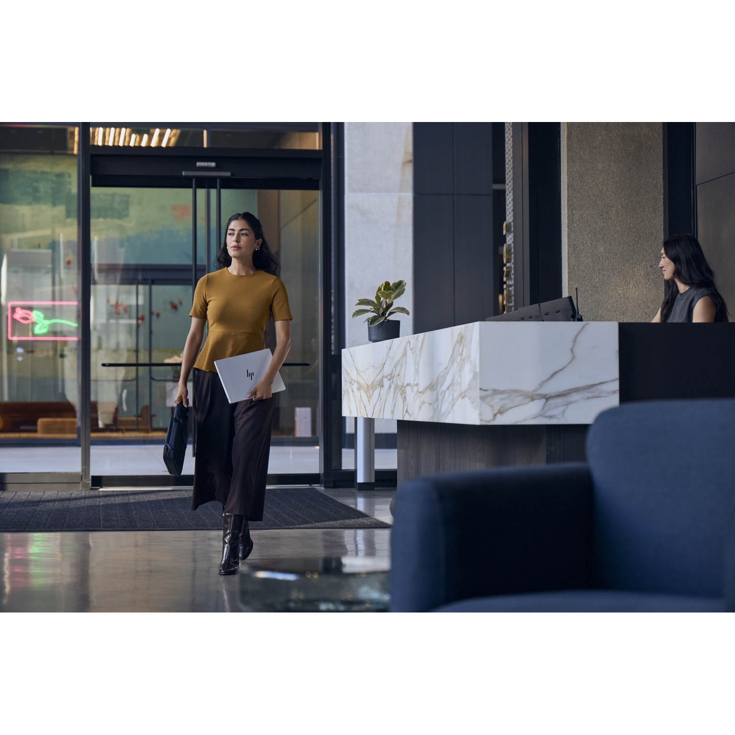 Een vrouw in een geel topje en bruine rok loopt door een moderne lobby terwijl een andere vrouw bij de receptie zit.