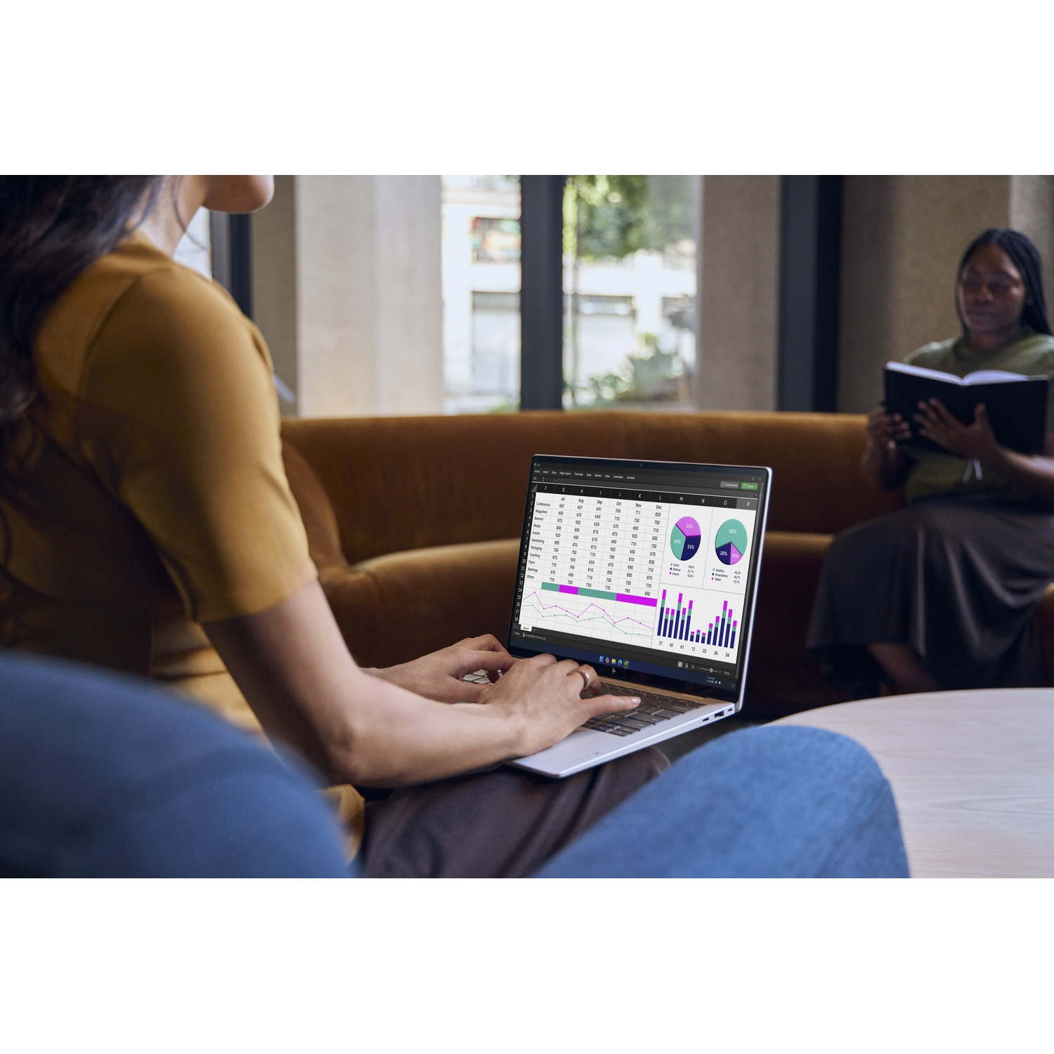 Een vrouw werkt op een laptop met datagrafieken in een helder kantoor, met iemand die leest op de achtergrond.