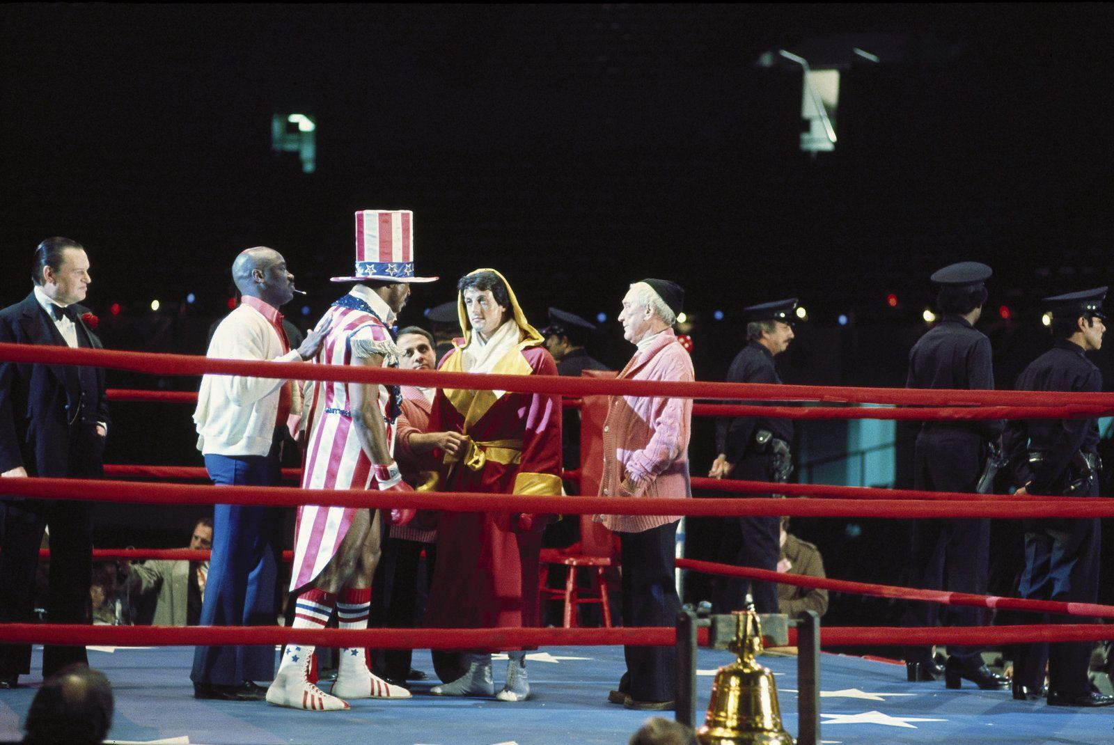 Boxer im Ring mit Männern im patriotischen Outfit und Mantel, im Hintergrund Polizisten und eine Glocke.