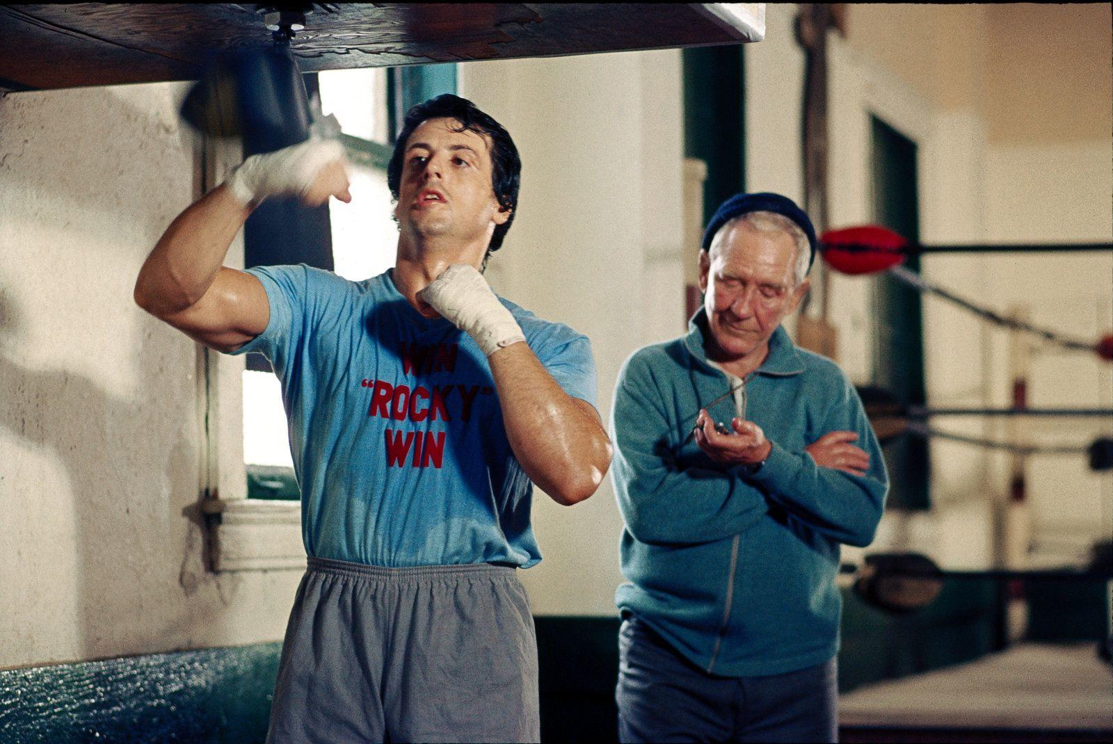 Rocky Balboa boxt gegen einen Boxsack, während sein Trainer zusieht.