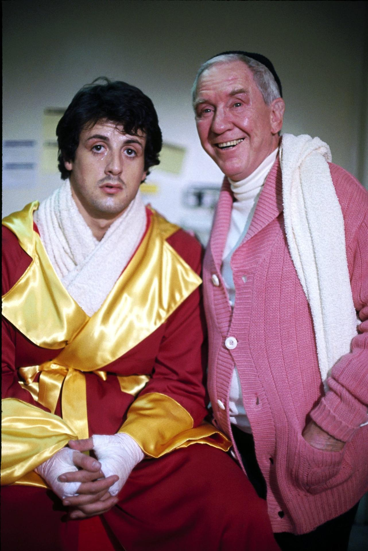 Sylvester Stallone in rotem Mantel und Burgess Meredith in rosa Strickjacke lächeln in die Kamera.