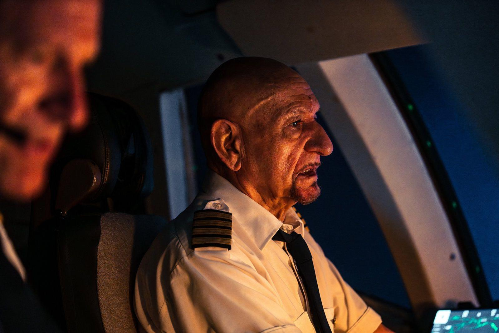 Ein älterer, kahlköpfiger Pilot in weißer Uniform sitzt im Flugzeugcockpit.