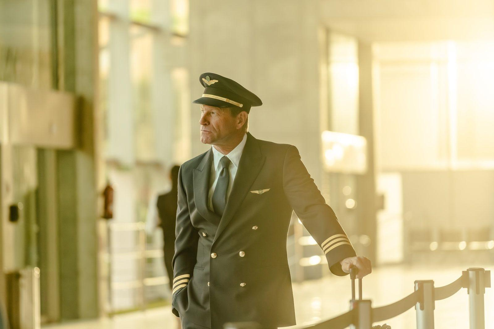 Ein Flugkapitän in Uniform steht in einem Flughafenterminal, hält Gepäck und schaut nach vorne.