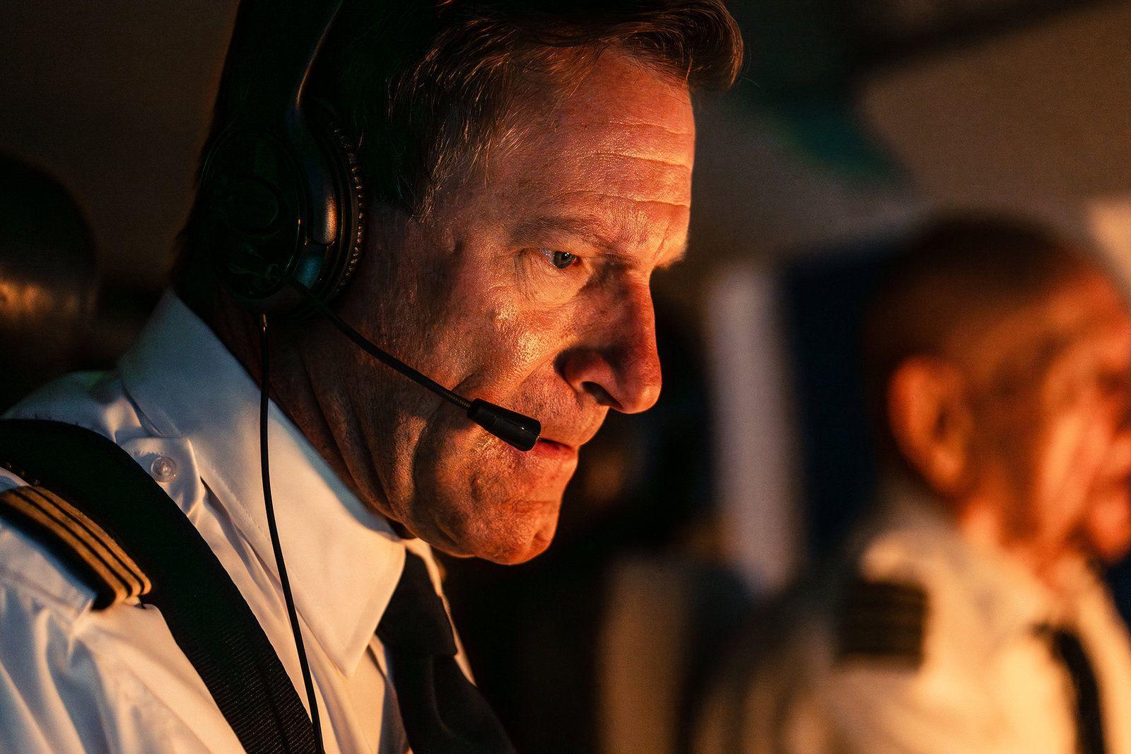 Ein Pilot in Uniform trägt ein Headset im beleuchteten Cockpit, eine andere Person sitzt im Hintergrund.