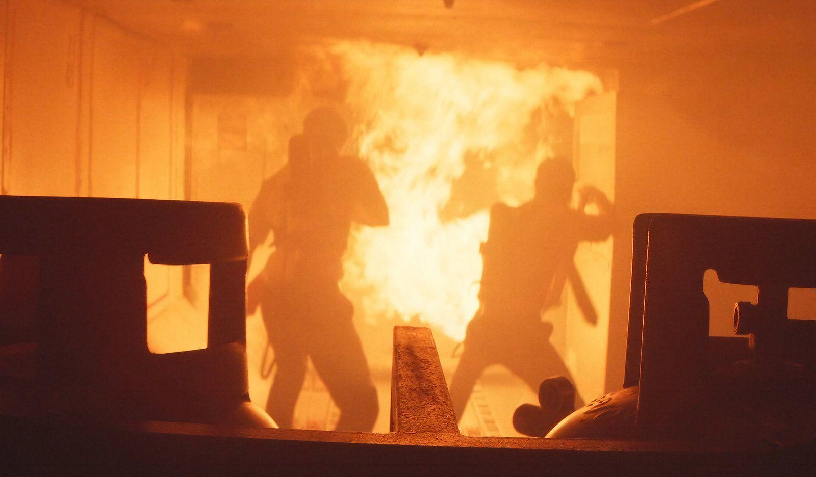 Zwei Feuerwehrleute als Silhouetten vor einer Wand aus hellem orangen Feuer in einem Raum, eingerahmt von dunklen Vordergrundobjekten.