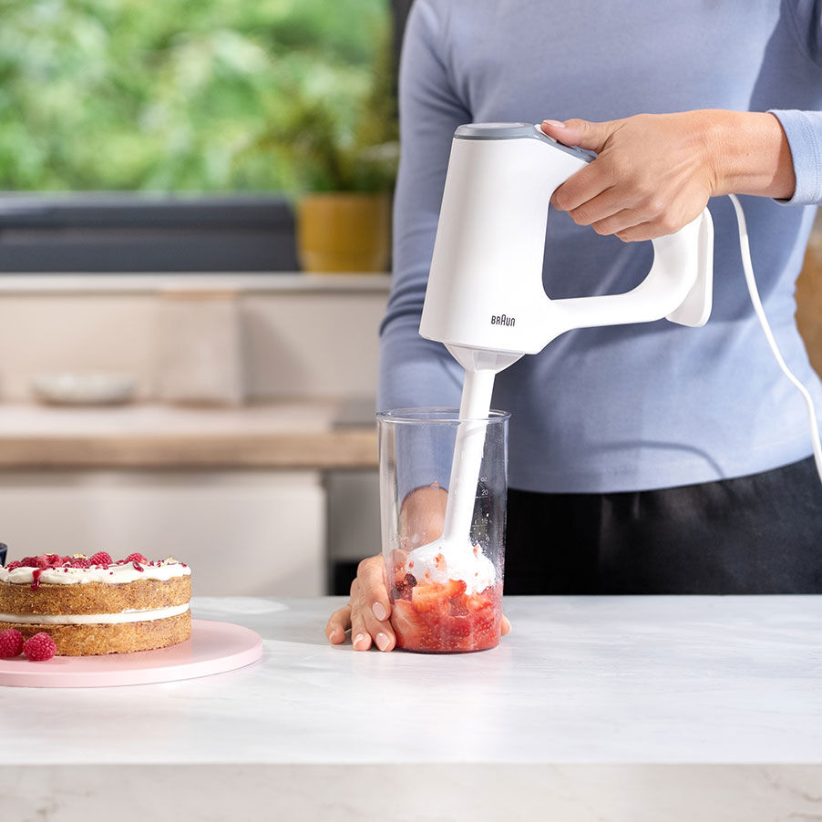Eine Person benutzt einen weißen Stabmixer, um Erdbeeren in einem Becher zu pürieren, daneben ein Himbeerkuchen.