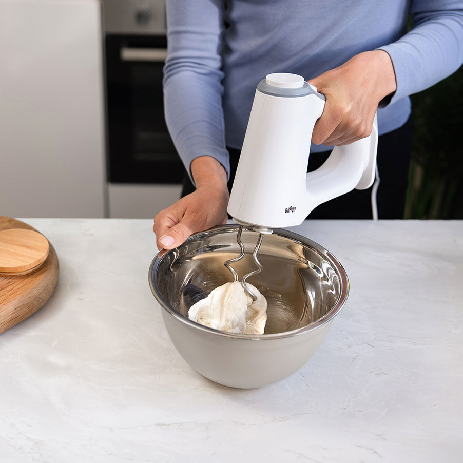 Eine Person benutzt einen weißen Handmixer in einer Edelstahlschüssel auf der Küchentheke.