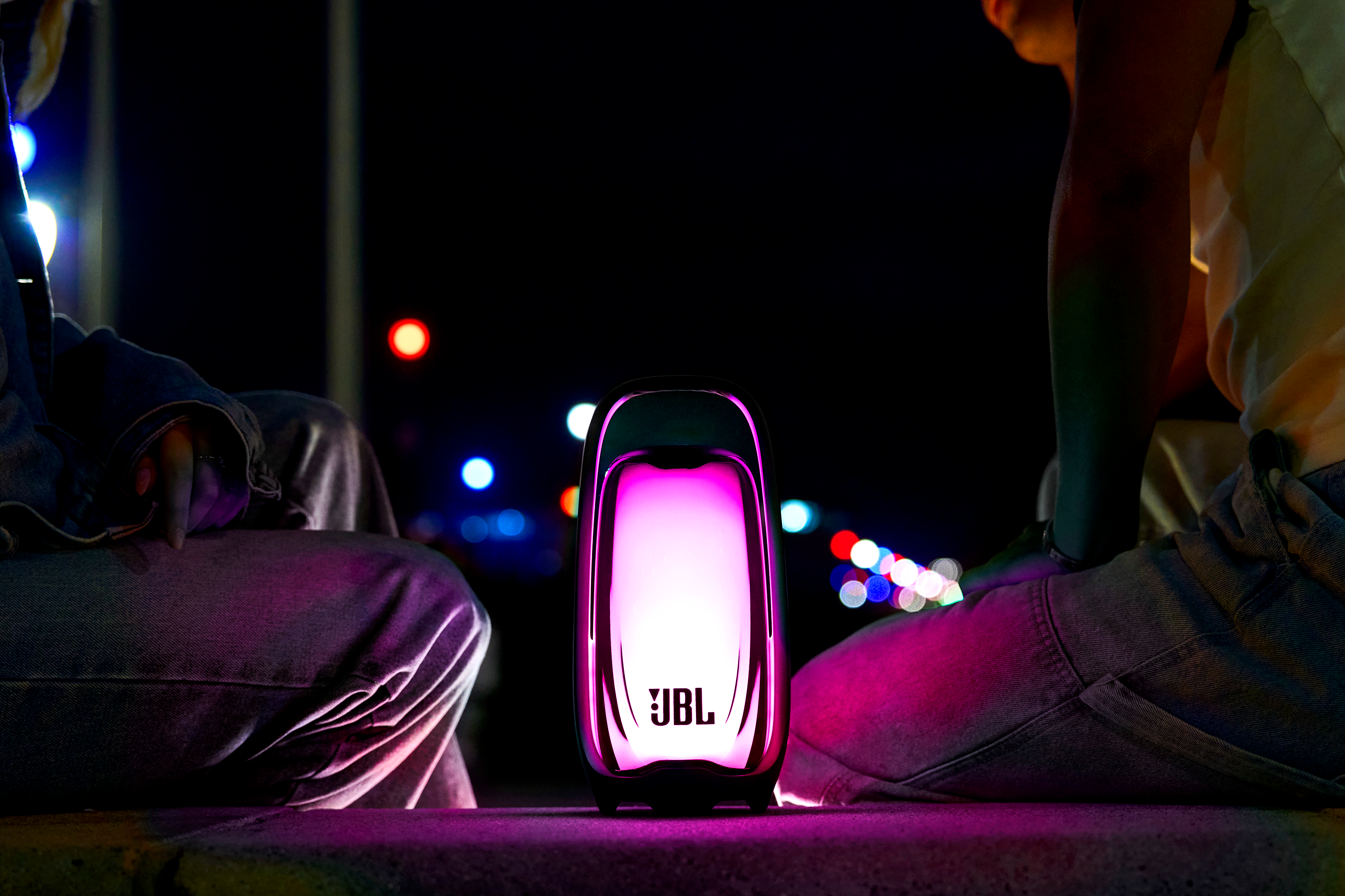 Haut-parleur portable JBL éclairé au sol la nuit avec deux personnes assises en arrière-plan.