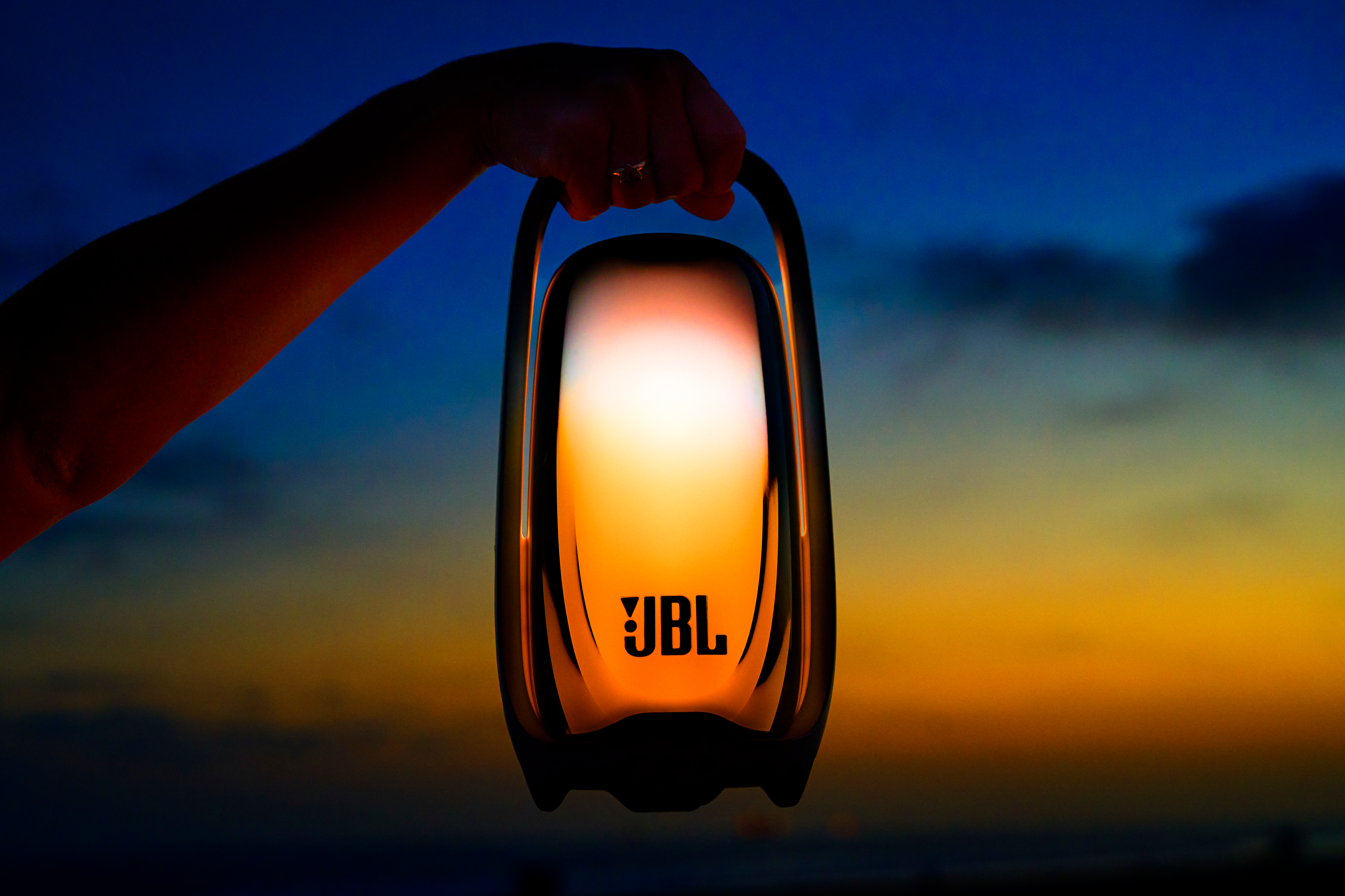 Une main tient une enceinte JBL éclairée sur un fond de coucher de soleil.