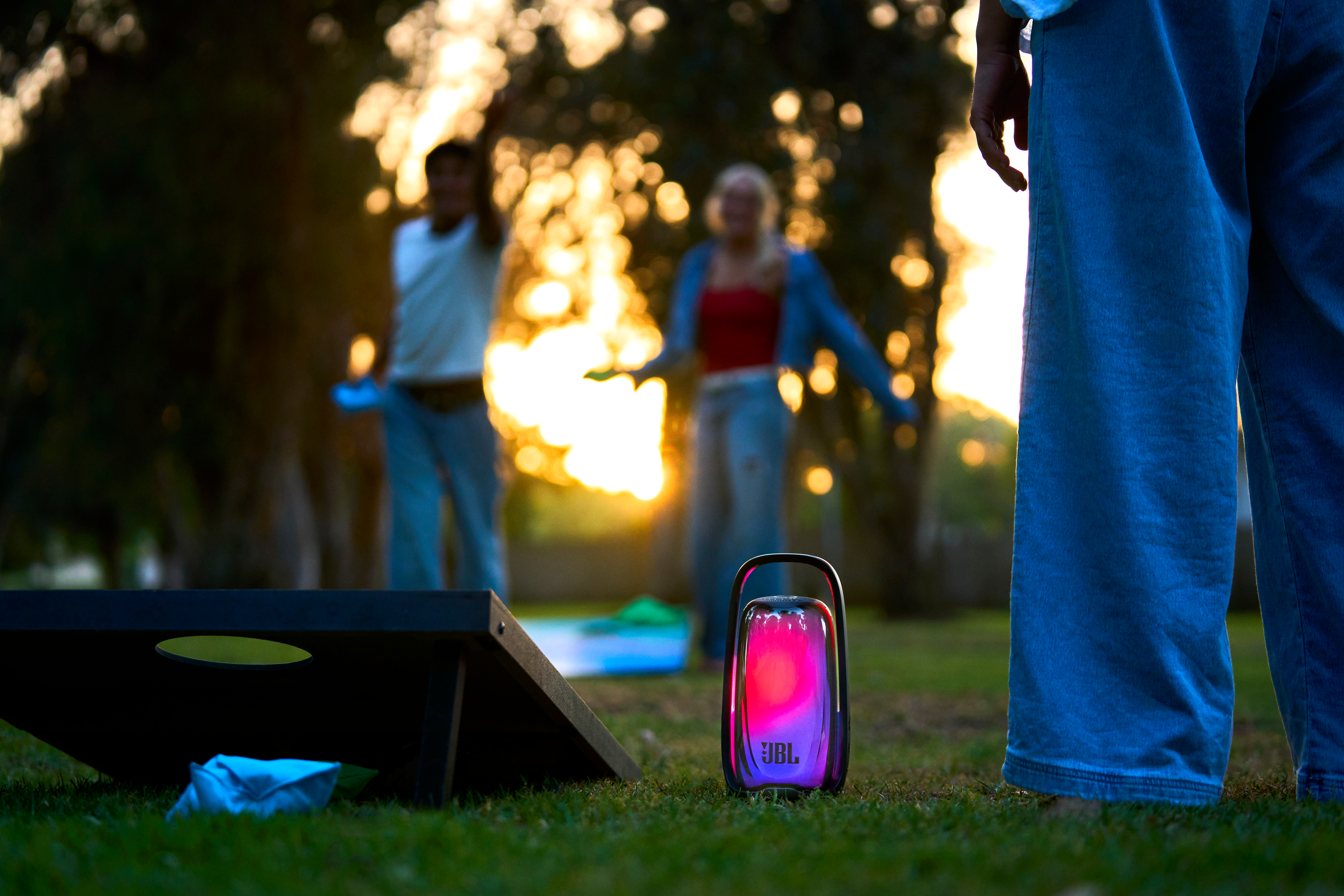 Une enceinte JBL lumineuse repose sur l'herbe d'un parc, avec des personnes floues en arrière-plan.