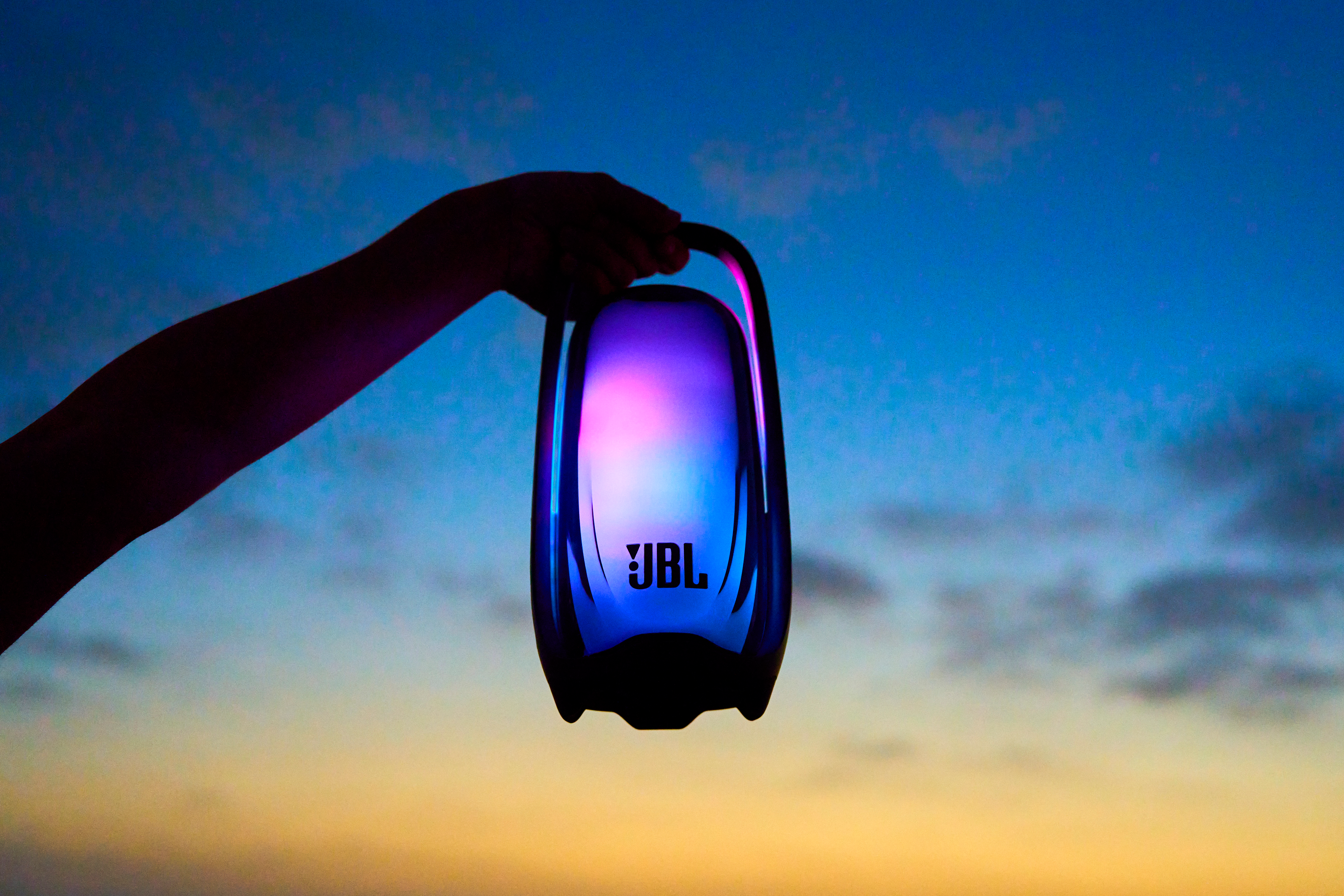 Une main tient un haut-parleur JBL éclairé sur fond de ciel du soir bleu et jaune.