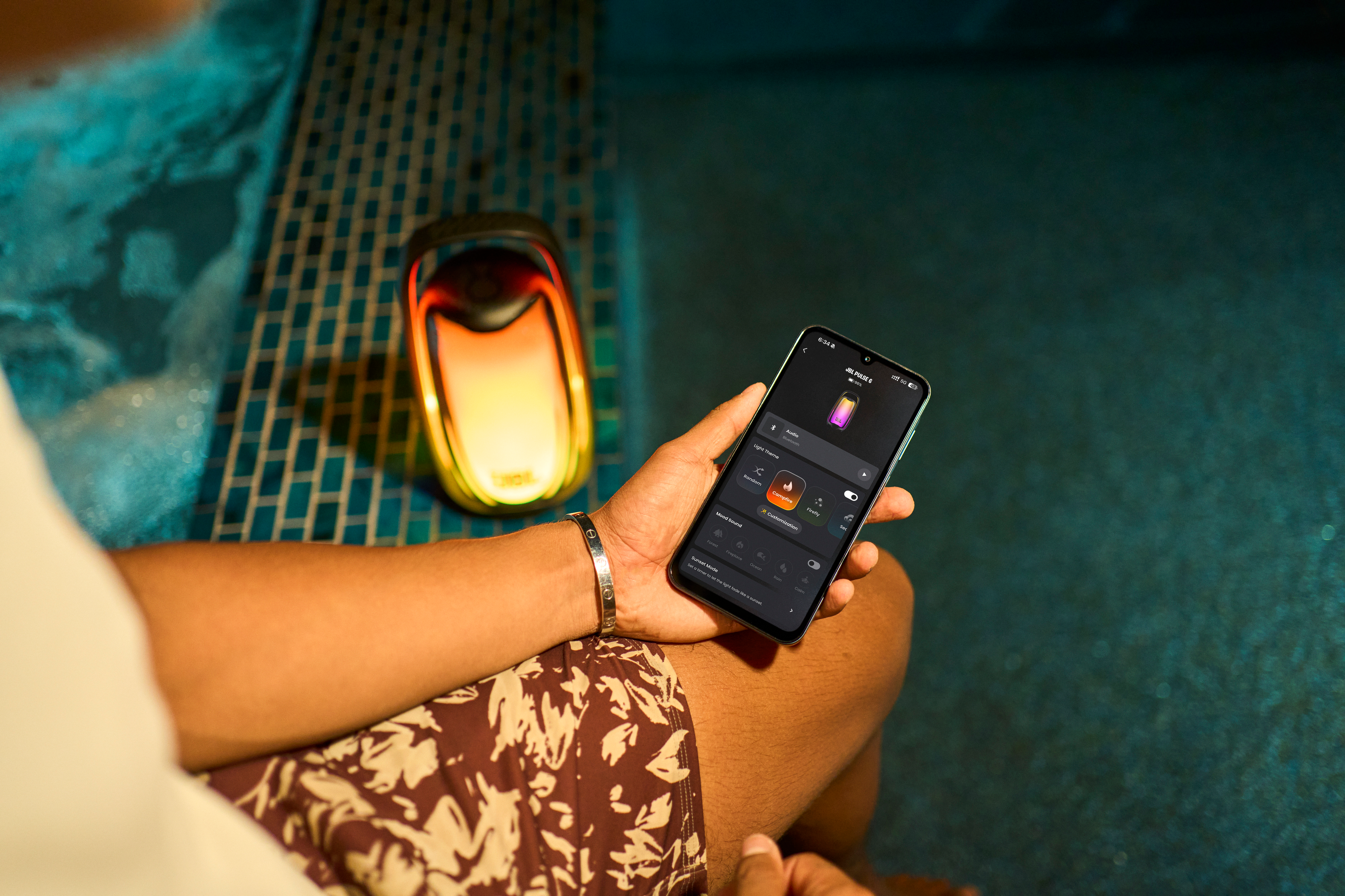Une personne assise au bord d'une piscine utilise une application mobile pour contrôler une enceinte lumineuse.
