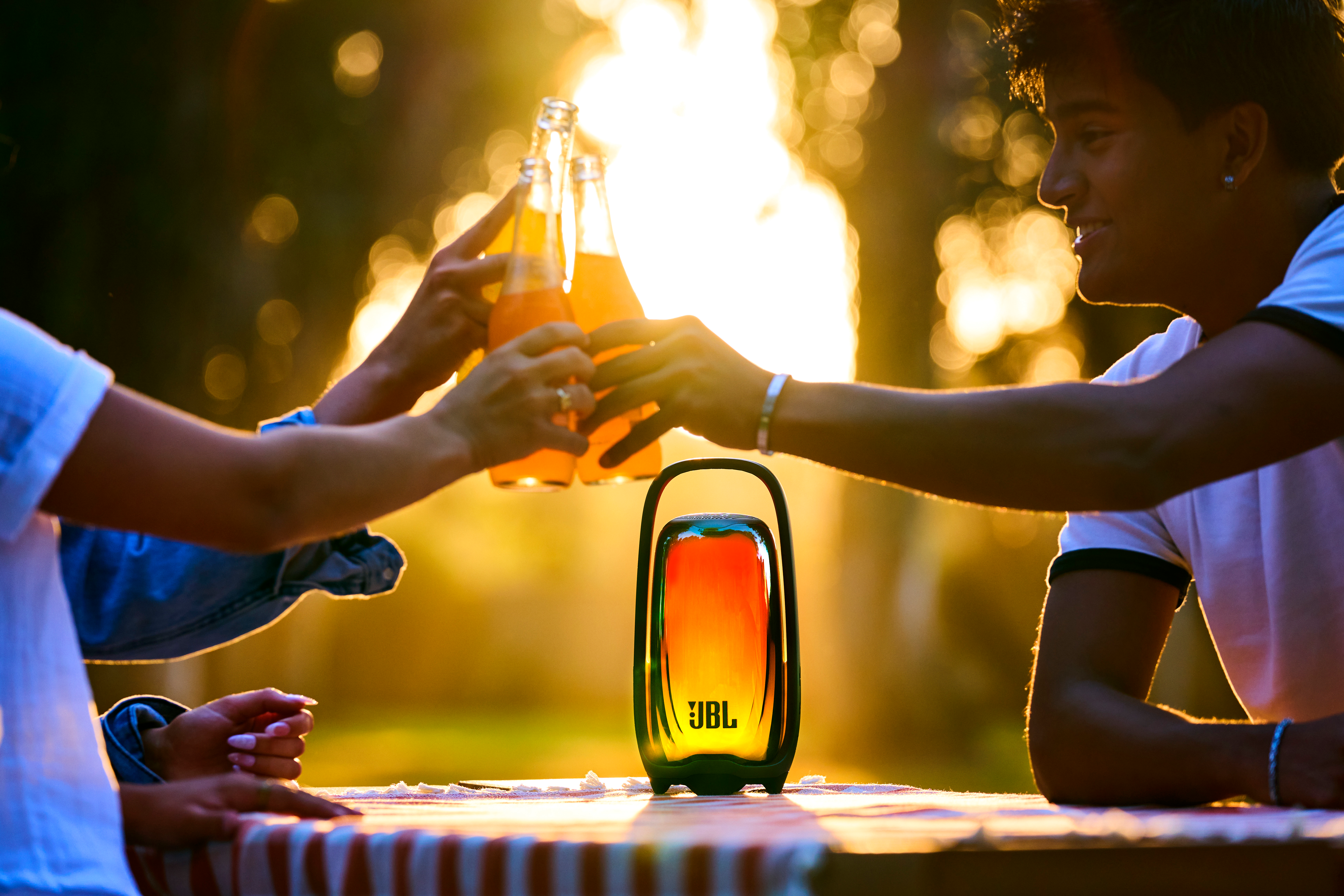 Des amis portent un toast avec des boissons au coucher du soleil, avec une enceinte JBL lumineuse sur la table.
