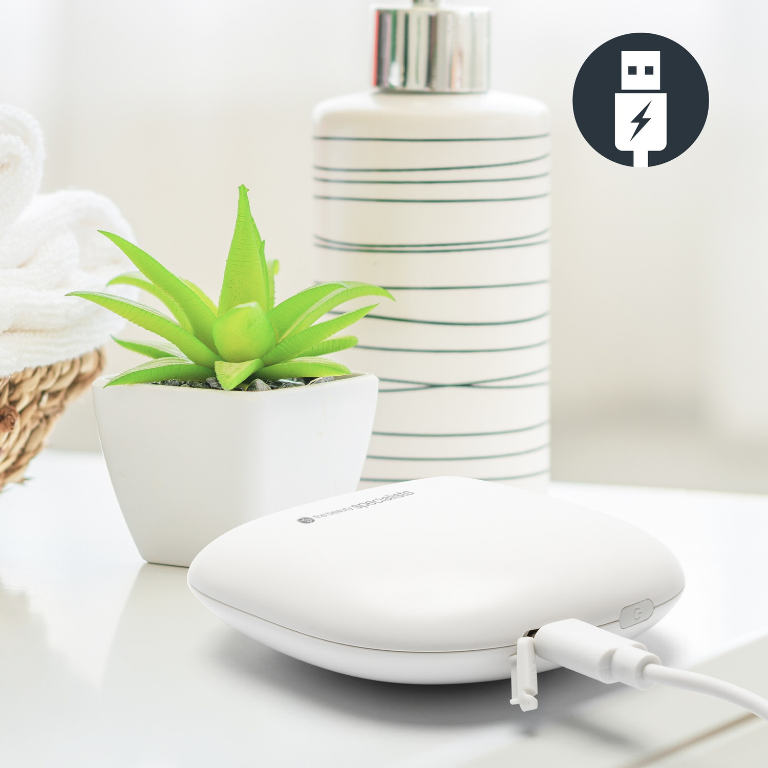 Un dispositivo di ricarica bianco con cavo su un tavolo vicino a una pianta e a un dispenser.