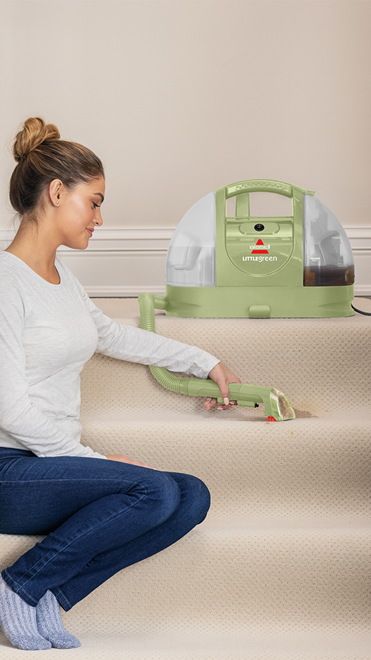 Bir kadın merdivenlerde diz çökerek yeşil bir Bissell Little Green elektrikli süpürgeyle halıyı temizliyor.