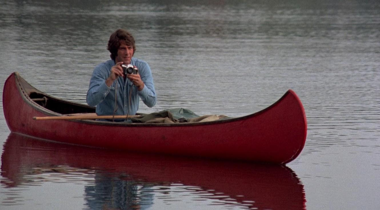 Ein Mann im blauen Hemd sitzt in einem roten Kanu auf dem Wasser und fotografiert mit einer kleinen Kamera.