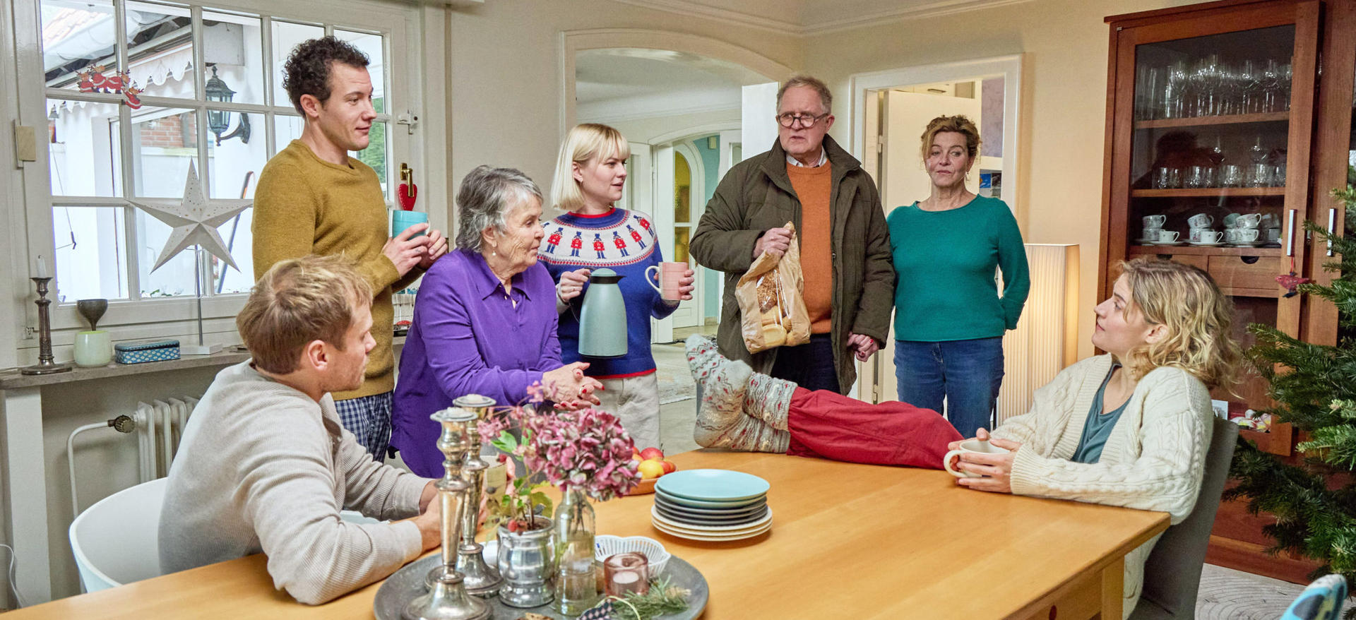 Eine Familie versammelt sich in einem hellen Esszimmer und unterhält sich an einem großen Holztisch mit Geschirr.
