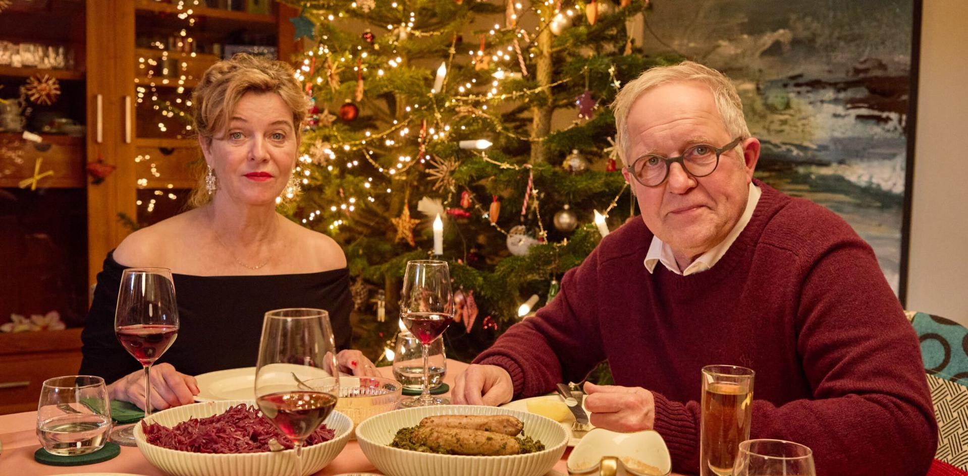 Ein Paar sitzt vor einem geschmückten Weihnachtsbaum beim Abendessen mit Würstchen und Rotkohl.