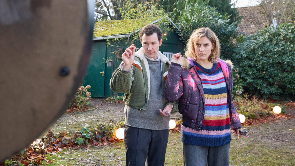 Ein Mann und eine Frau stehen im Garten und halten jeweils ein kleines Objekt hoch, im Hintergrund ein grüner Schuppen.