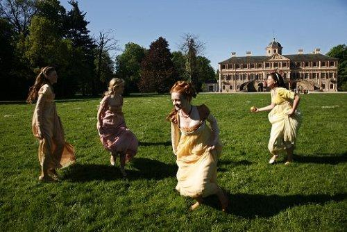 Vier Frauen in historischen Kleidern laufen fröhlich über einen grünen Rasen vor einem großen Schloss.