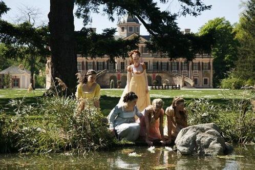 Frauen in historischen Kleidern versammeln sich im Park vor einem prunkvollen Herrenhaus an einem Teich.