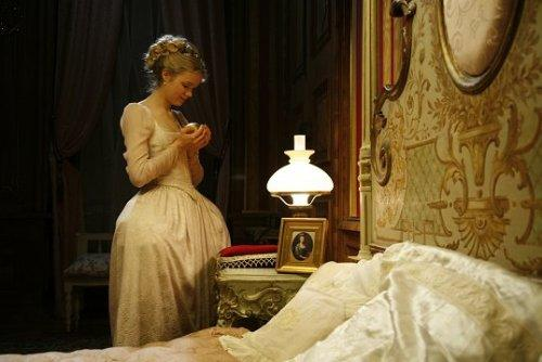 Eine Frau im historischen Kleid steht im gedimmten Schlafzimmer neben einem verzierten Bett und einer beleuchteten Lampe.