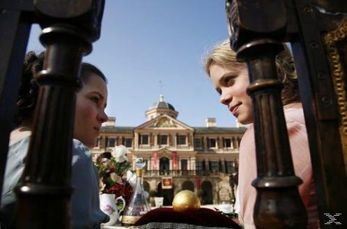 Zwei Frauen sitzen im Freien vor einem großen Schloss, eingerahmt von dunklen Holzpfeilern.