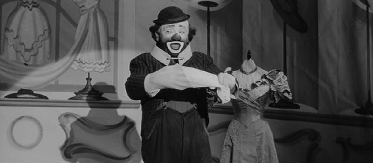 Ein Clown mit Hut und weißem Kragen steht in einem Raum mit Kleiderpuppen.