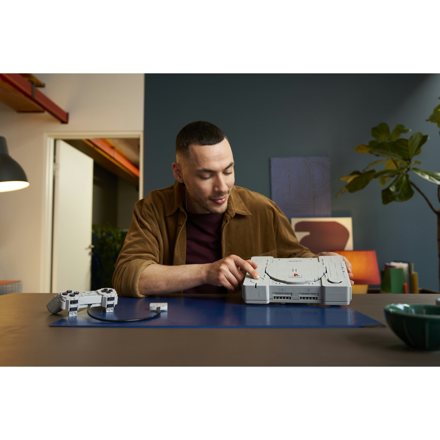 Un uomo con una camicia marrone siede a un tavolo costruendo un modello Lego della PlayStation.