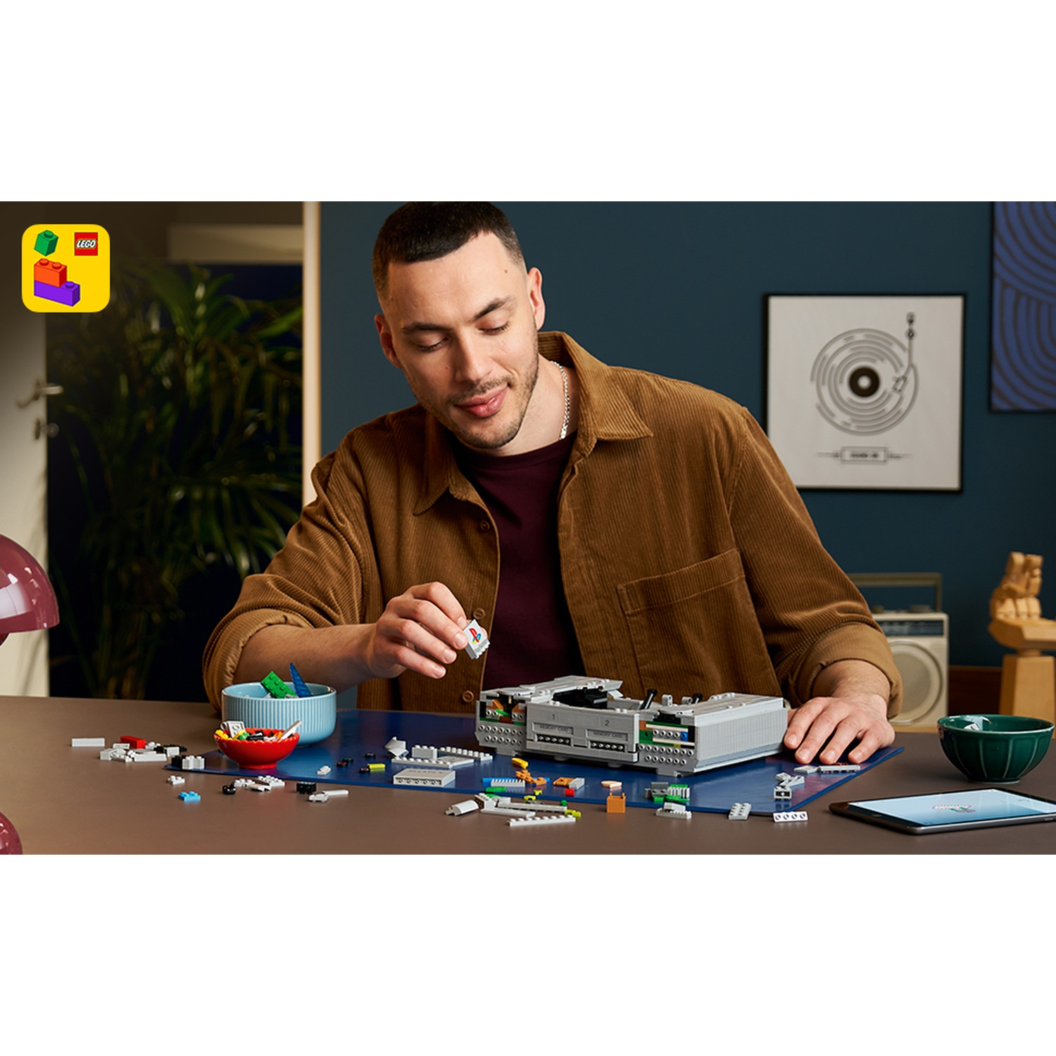 Un uomo costruisce un modello Lego grigio di una console di gioco su un tavolo di legno.